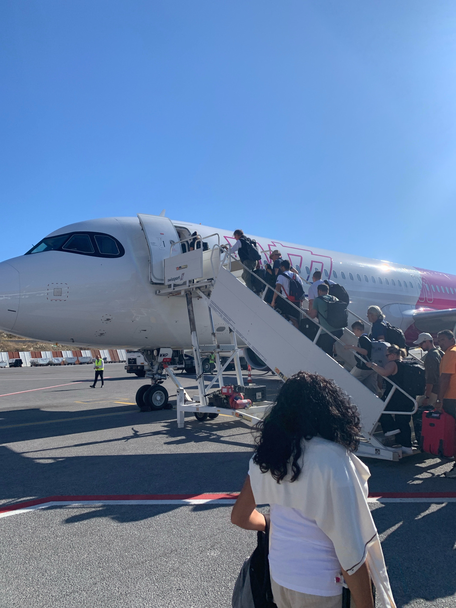 This is a photograph taken on the tarmac at Skiathos Airport (JSI), Greece. The image shows passengers boarding a white airplane with a pink "Wizz Air" logo on its fuselage. A mobile staircase is attached to the aircraft's main door, and a stream of people are ascending it, carrying luggage. In the foreground, a woman with dark, curly hair is visible from behind, wearing a white tank top and beige shorts, with a white shawl draped over her shoulders. She is facing away from the camera and towards the airplane. Behind her, more passengers are seen on the stairs and the tarmac. Several airport vehicles and equipment are present in the background, including service trucks and baggage carts. The scene is under a clear blue sky, suggesting a sunny day. The ground is asphalt, marked with yellow and red lines.