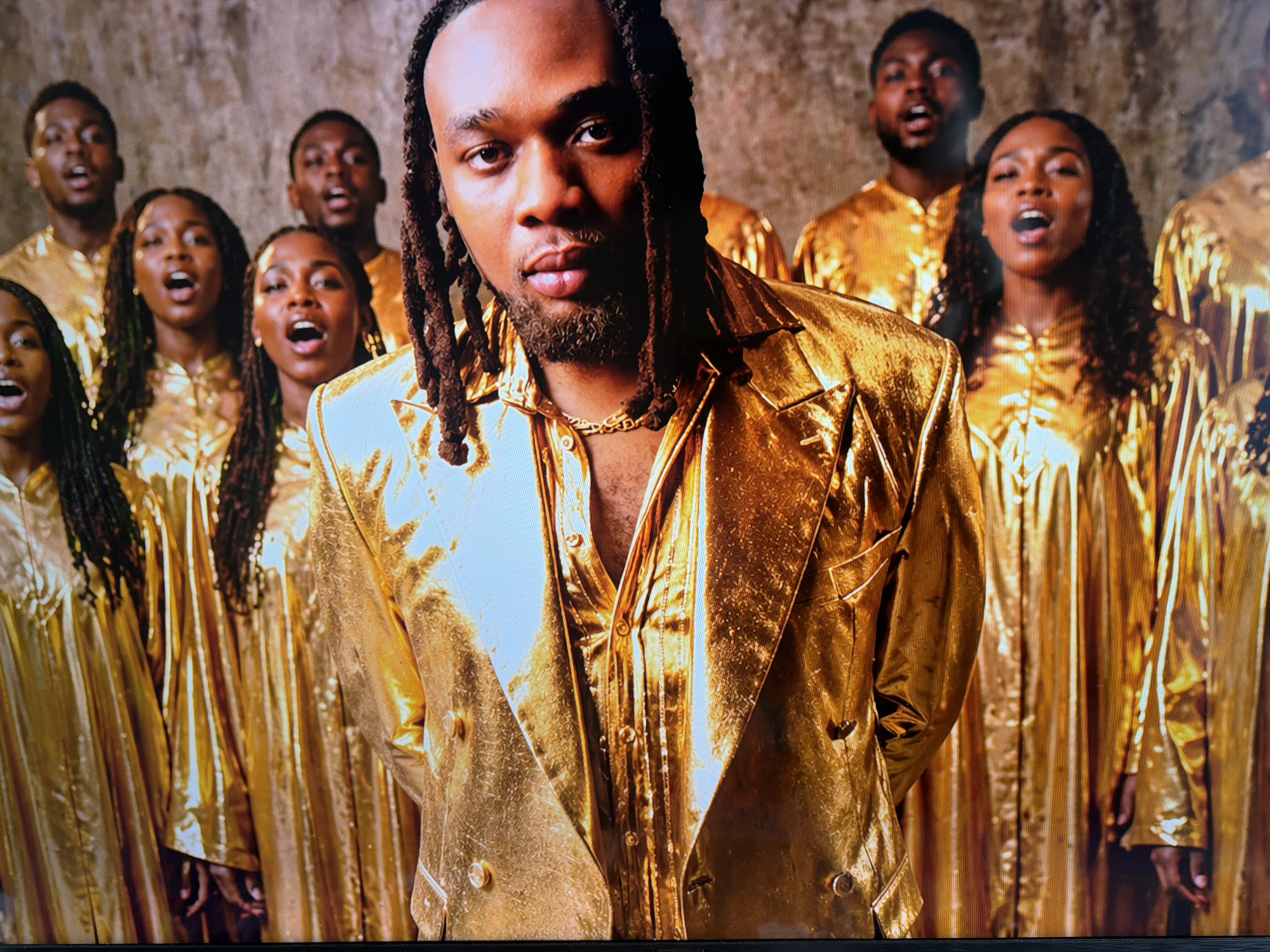 This image captures a group of Black individuals, primarily young adults, positioned against a textured, possibly stone or concrete, background. In the foreground, a man with long dreadlocks and a beard is centrally featured, looking directly at the viewer with an intense and focused expression. He is dressed in a striking, shiny metallic gold jacket over a gold button-down shirt, complemented by a gold chain around his neck.
Behind him, several other individuals are arranged in rows, creating the impression of a choir or musical group. Their mouths are open, indicating they are singing or performing with emotion and concentration. All the background figures are uniformly clad in lustrous, metallic gold robes or gowns, which prominently reflect the strong lighting. Some of the women in the background also have long, braided hair or dreadlocks.
The overall scene conveys a sense of a powerful musical performance or a staged portrait, possibly for an album cover or promotional material. The rich, golden attire and the expressions of the singers suggest an uplifting or spiritual atmosphere. The strong, artificial lighting highlights the reflective quality of the garments, making them a central visual element. No text is visible in the image. The setting is indoors, with no discernible outdoor weather or time of day cues, and the location context is Cape Town, South Africa.