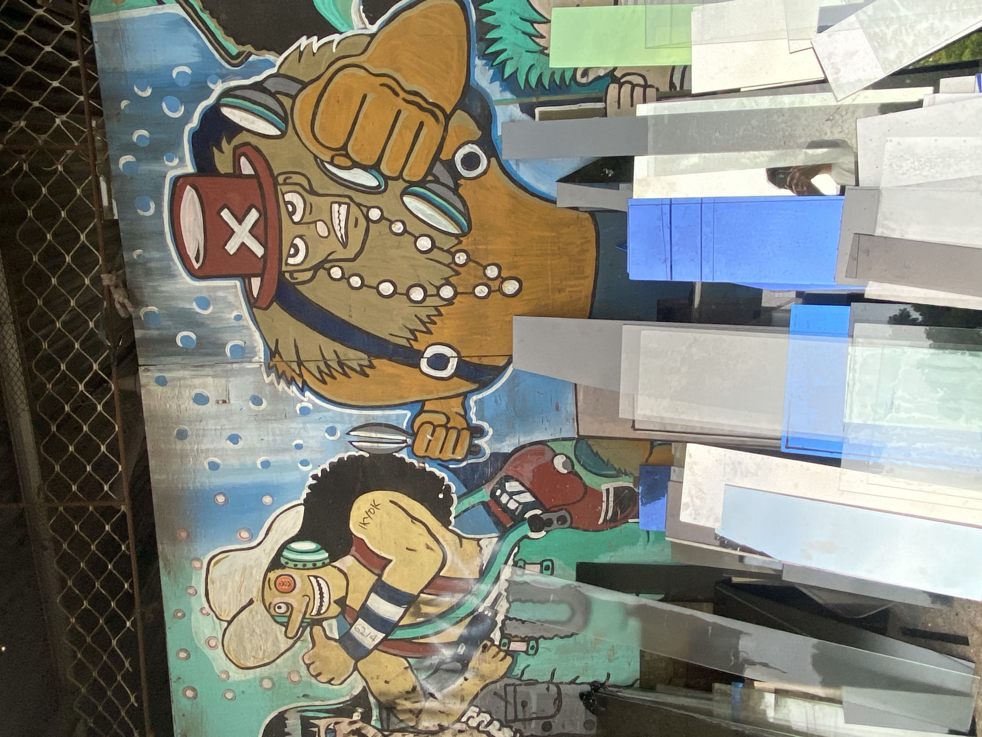 The image shows a graffiti mural of characters from the anime series One Piece, specifically Usopp and Chopper, on a weathered wooden wall. The mural is located in Davao City, Philippines, as indicated by the graffiti text "IKYOK" which is a reference to the local Davao City slang. In the foreground, a pile of various colored glass sheets is stacked. The glass reflects the graffiti and the surrounding environment. The upper portion of the image shows a metal mesh fence. The overall scene appears to be an outdoor urban setting, possibly a workshop or storage area.