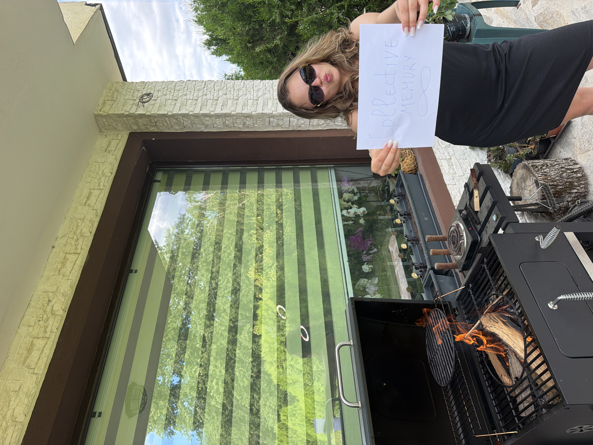 A young woman with long, wavy brown hair and sunglasses is standing on a patio in front of a large window. She is wearing a black dress and holding a white piece of paper with the words "Collective Memory" and an infinity symbol drawn on it. A charcoal grill with burning wood is to her left. The patio is paved with light-colored stones and surrounded by greenery. In the background, there are trees and a cloudy sky. The architecture of the building suggests a suburban or rural setting. The image was taken in Priseltsi, Bulgaria.