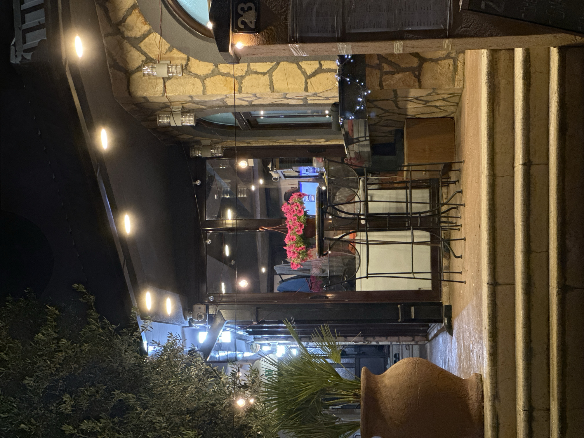 The image depicts an outdoor dining area in Donji Pobori, Montenegro, likely a restaurant. The area is adjacent to a stone building with the number "23" displayed. Outdoor seating is visible, featuring metal tables and chairs. Potted plants, including flowering plants and a palm, decorate the area. String lights illuminate the space, and the scene appears to be taken at night. A set of stairs runs along one side of the dining area.