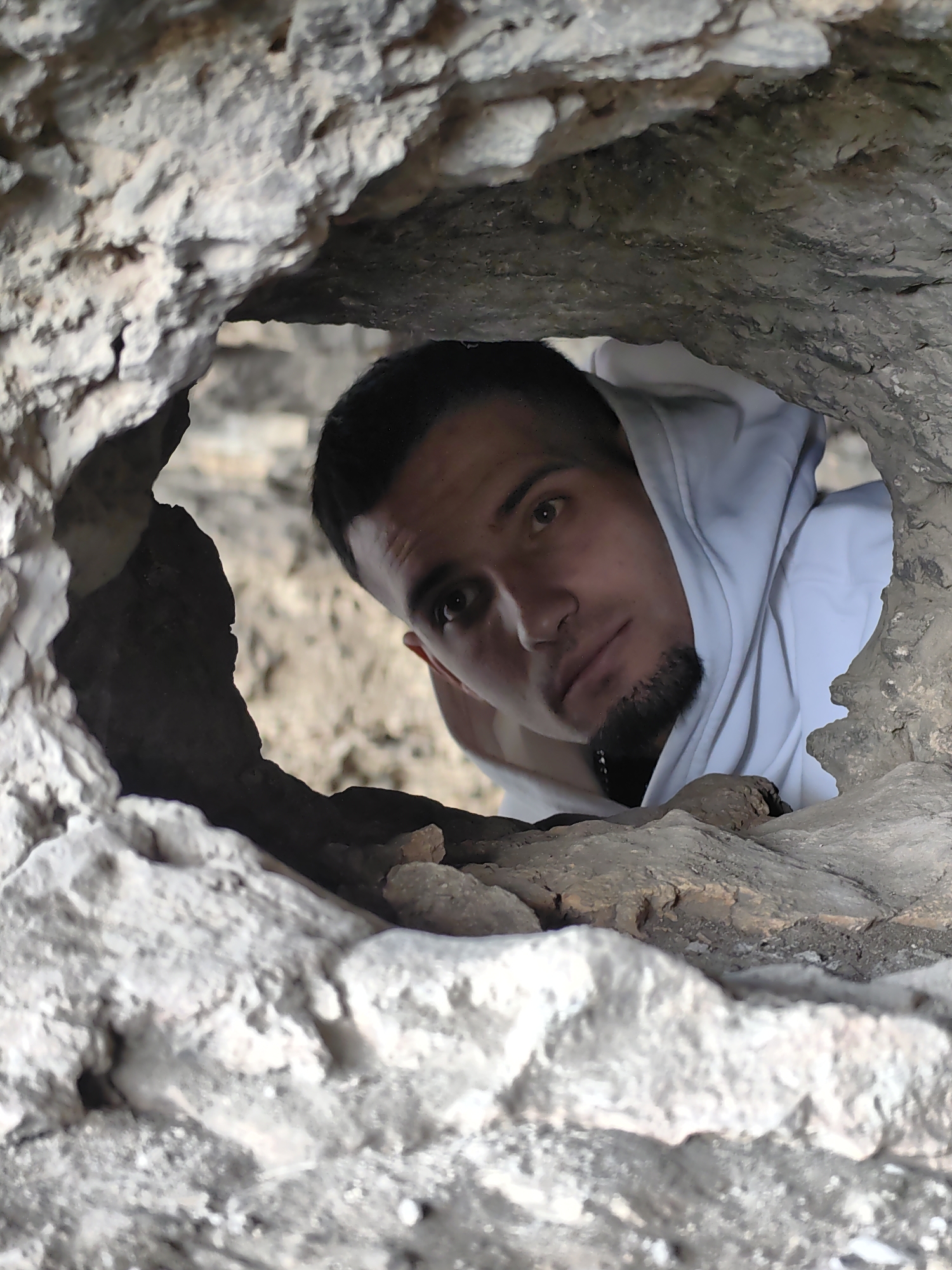 A young man wearing a white hooded sweatshirt is looking through a hole in a rough, light-colored rock formation. The location is potentially in or near the city of Marash, Bulgaria, though this cannot be verified solely from the image. The hole is irregular in shape, with jagged edges. The man's face is in focus, and his expression appears contemplative. Part of the sweatshirt is visible around his head and shoulders. The surrounding rock is textured and shows signs of weathering. The background is blurry.