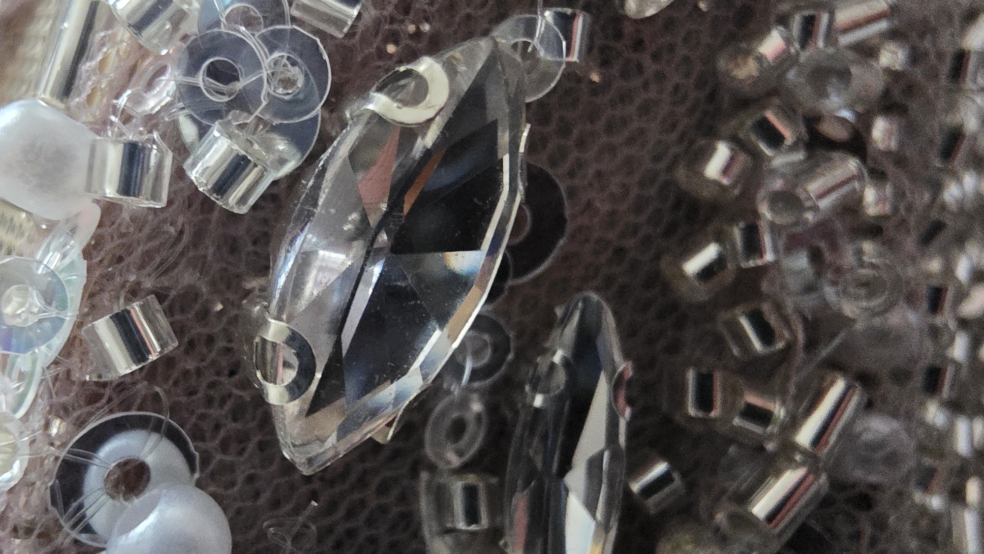 The image shows a close-up of decorative embellishments on a textured surface. Dominating the scene are several large, clear, faceted crystals or gemstones, reflecting light and creating internal refractions. Accompanying these are numerous smaller, cylindrical silver beads and clear, flat sequins with central holes. A single, round, opaque white pearl is also visible. The background appears to be a soft, textured fabric in a muted brownish-gray tone. The focus is sharp on the foreground elements, particularly the large crystals, while the background has a softer focus. There is no indication in the image of the location of Blagoevgrad, Bulgaria. The scene depicts inanimate objects; there are no people or actions visible. The interaction is of the light with the various materials, creating highlights and shadows.