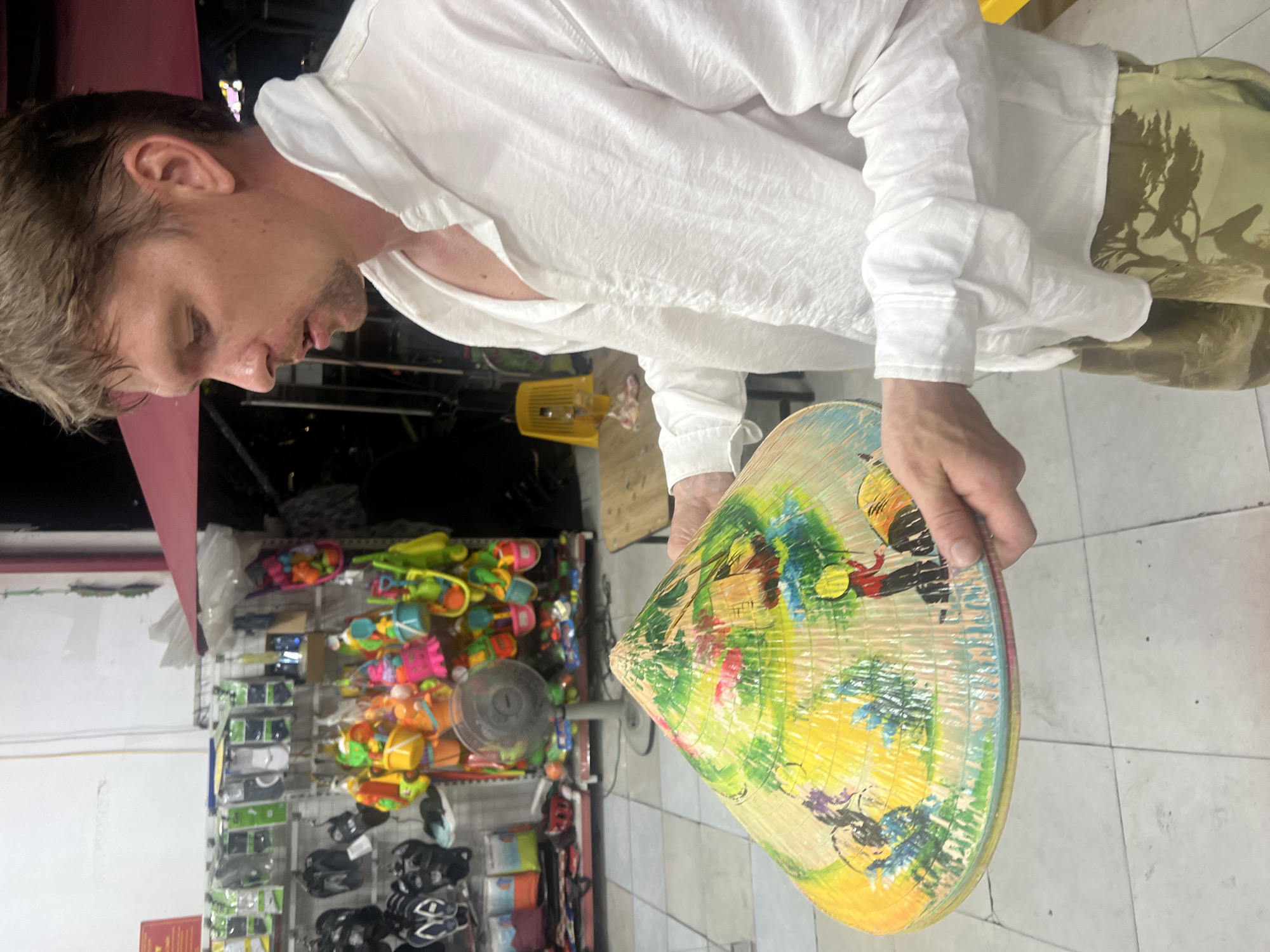A fair-skinned man with short brown hair is standing in what appears to be a shop in Da Nang, Vietnam. He is wearing a loose-fitting white shirt and camouflage shorts. In his hands, he holds a conical hat, which is decorated with colorful paintings. The hat depicts a scene with what looks like people and a landscape. Behind him, shelves are stocked with various items, including what seem to be flip-flops and beach toys. A small electric fan is visible on a shelf in the background. The floor is tiled.