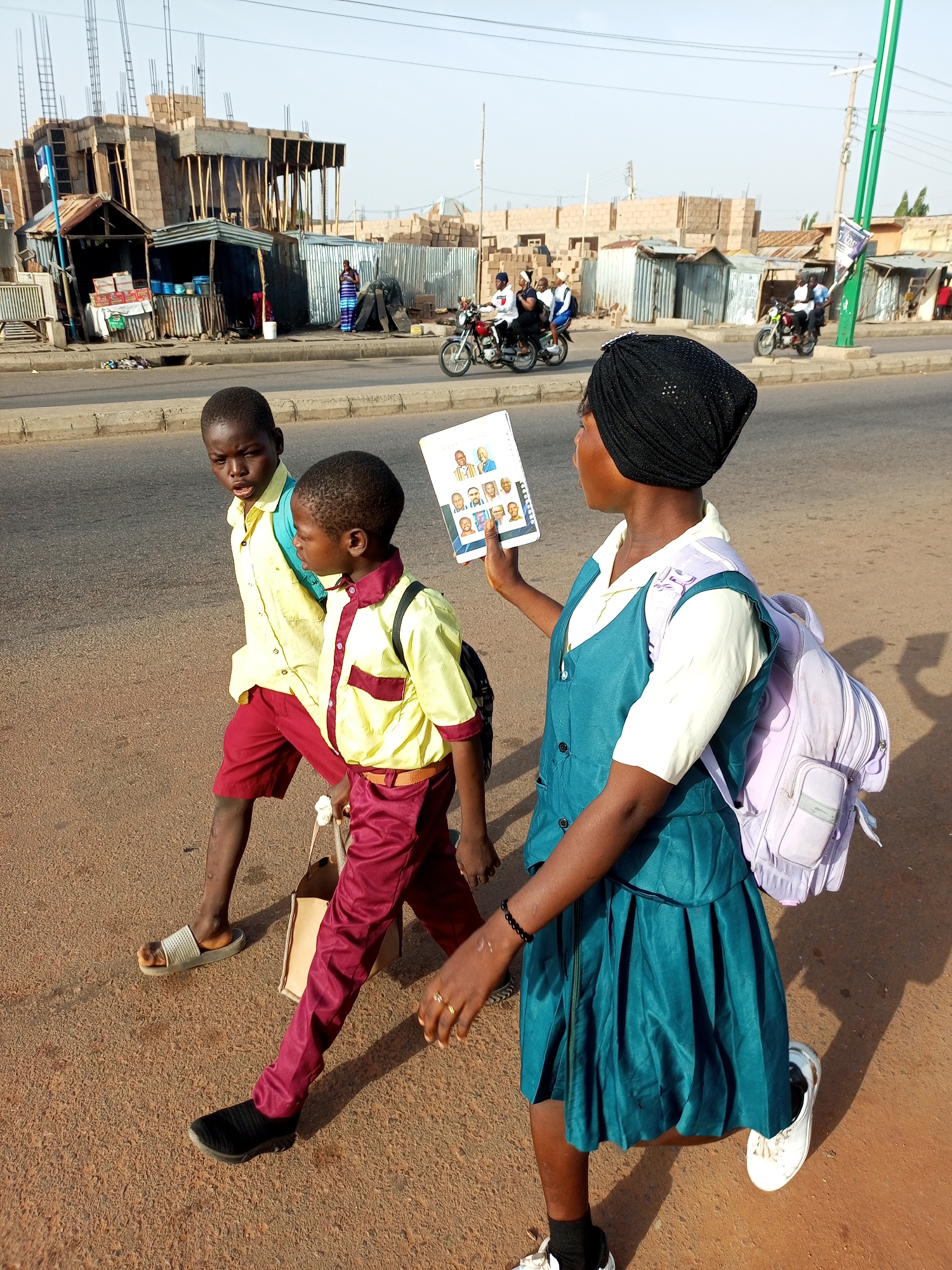 In Tempure, Nigeria, three children in school uniforms walk along a dusty road next to a busy street. The two boys in front wear yellow shirts with maroon shorts and red pants respectively, carrying backpacks. The girl behind them, wearing a turquoise pinafore over a yellow shirt and a black headscarf, holds a booklet with portraits. In the background, construction is underway on a building, and several motorcycles are visible on the road, some with riders. Small market stalls line the side of the street, and overhead utility poles are present. The scene is illuminated by natural daylight.