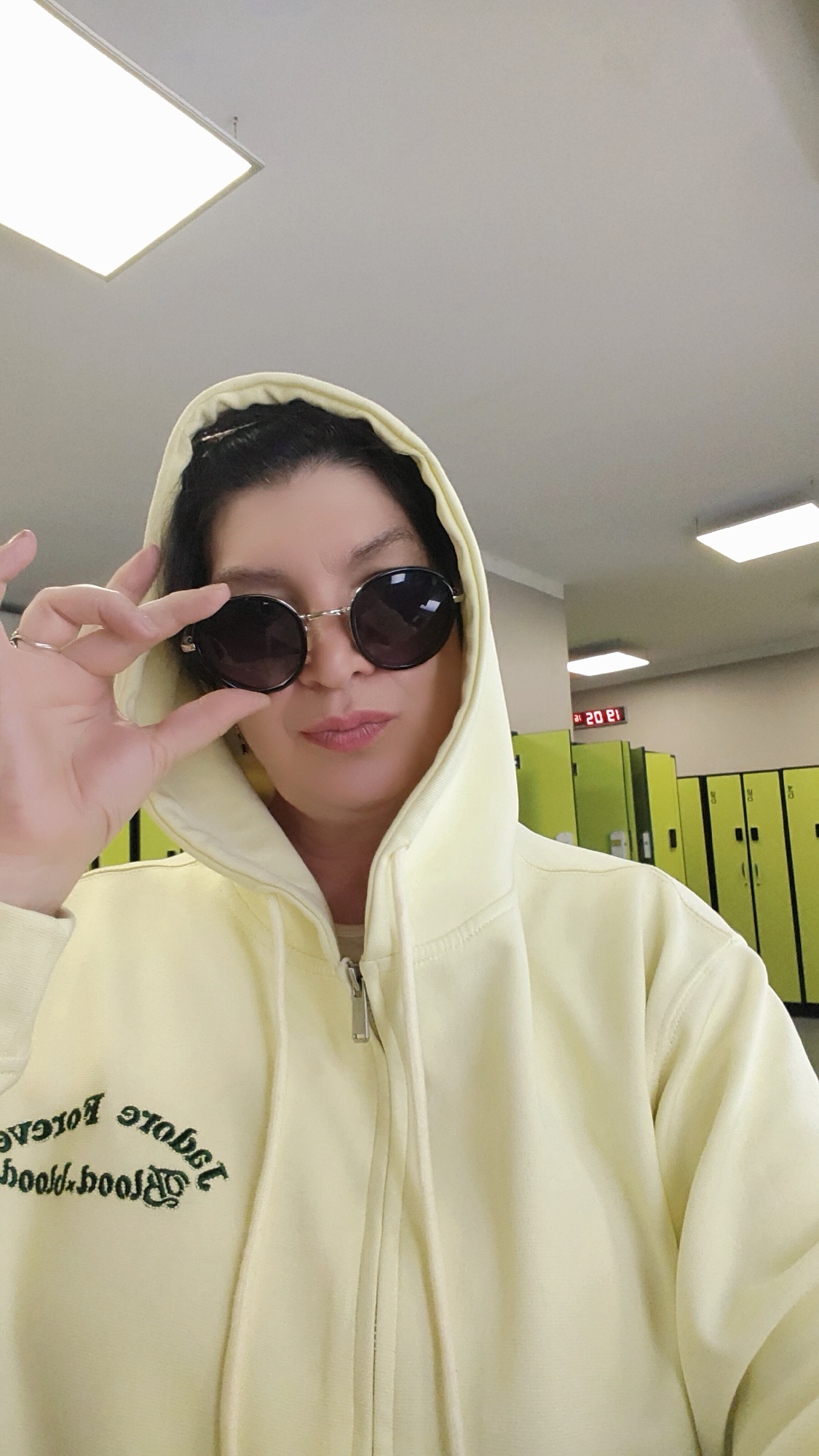 A woman in a light yellow hooded jacket is posing indoors, holding her sunglasses. Behind her are rows of green lockers and an electronic clock that reads 13:02. The name of the city is not discernable.