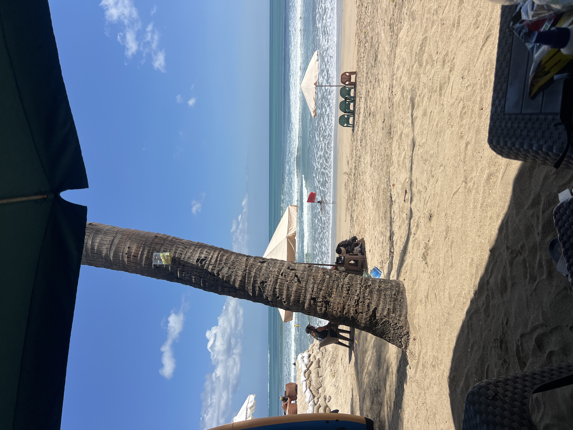 This is a photograph taken from a seated perspective, looking out towards a beach in Kuta, Indonesia. The foreground is dominated by the shadow of a large umbrella, obscuring the lower portion of the image. A portion of a woven chair is visible in the bottom left corner.
The midground features a tall, textured palm tree positioned to the left of center, with a small plastic cup containing yellow items attached to its trunk. Behind the palm tree, a beach with light brown sand stretches towards the ocean. Several beach chairs are visible, some clustered together under a light-colored umbrella, while others are spaced out. A red flag is planted in the sand to the left of the umbrella.
In the background, the ocean is a vibrant turquoise color, with gentle waves rolling towards the shore. The horizon is clearly defined against a bright blue sky dotted with scattered white clouds. Several people are visible on the beach and in the water, some appearing to be surfers. To the far left, a portion of a surfboard is visible. The overall impression is a sunny and relaxed beach scene.