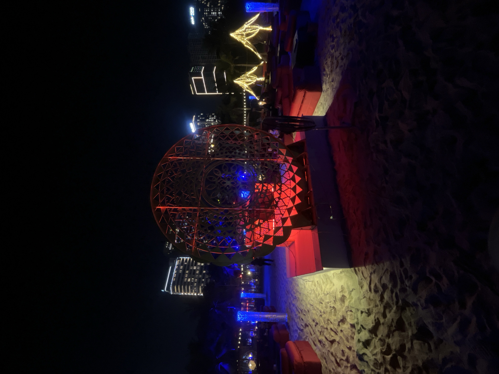 This image depicts a vibrant night scene at a beachside location in Đà Nẵng, Vietnam. The foreground consists of sandy ground, illuminated by various colored lights, casting shadows and highlighting its textured surface.
A large, prominent, spherical decorative structure, intricately designed with geometric patterns, stands on the left side of the frame. This sphere is internally lit with shifting red and blue lights, making it a focal point. To the right, several red lounge seating arrangements, possibly sofas or cushions, are placed directly on the sand, suggesting a casual yet stylish outdoor lounge area.
Further back, more decorative lighting elements are visible, including two abstract, golden-lit sculptures that resemble stylized plants or trees, adding to the festive atmosphere. Blue illuminated vertical structures or tables are also interspersed among the seating. In the background, against a dark night sky, several modern high-rise buildings are silhouetted, their architectural features outlined by white and yellow lights.
The time of day is clearly night, with the dark sky contrasting sharply with the multitude of artificial lights that illuminate the scene. The setting appears to be an upscale outdoor leisure or entertainment area, likely a beach club or resort lounge. While no specific activity or people are clearly depicted, the arrangement suggests a place for relaxation, socializing, or enjoying the evening ambiance. There is no visible text in the image.