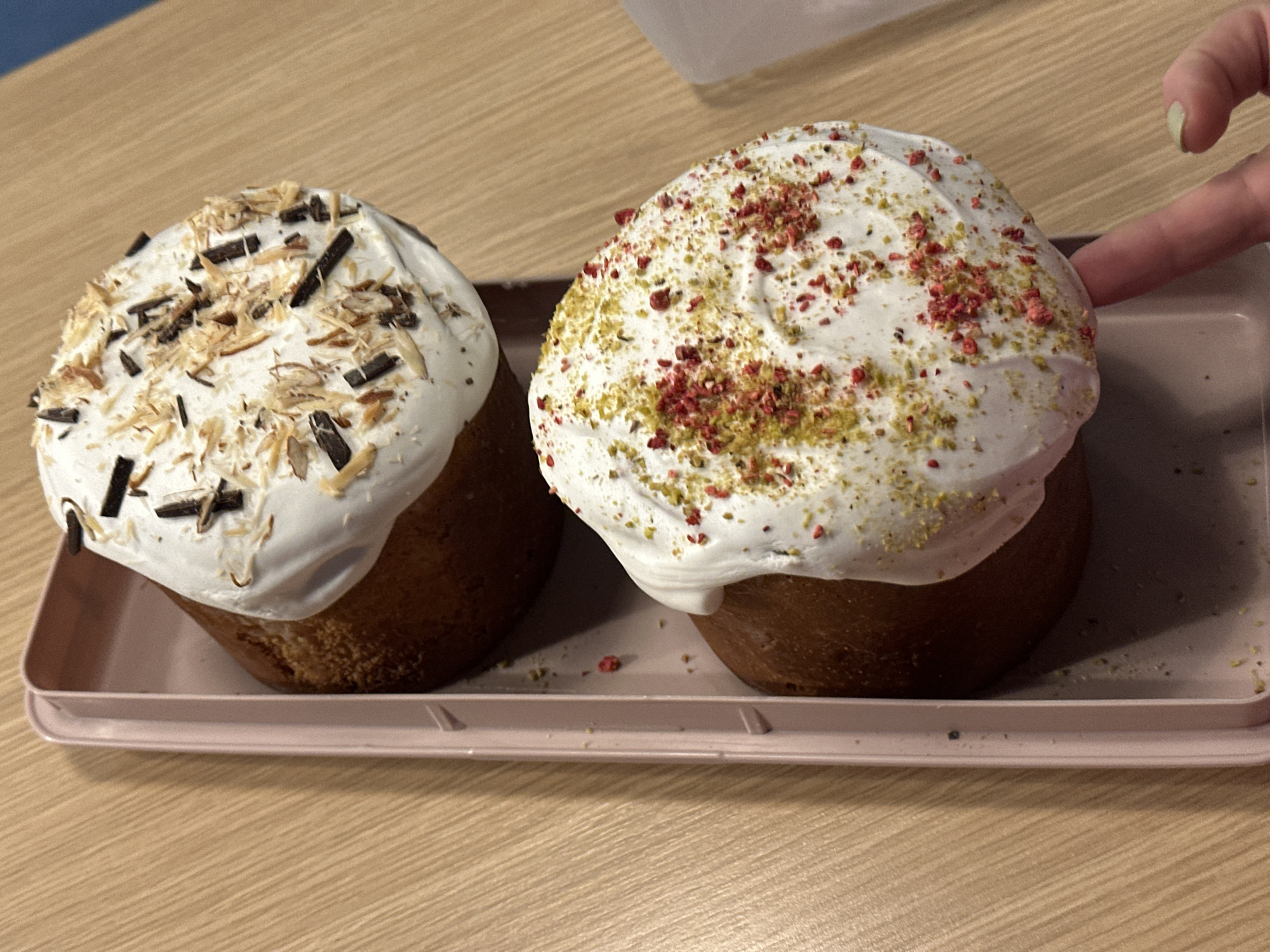 The image shows two Easter cakes (Kulich) presented on a light pink tray. The cake on the left is decorated with white icing, chocolate shavings, and flaked nuts. The cake on the right is similarly iced but adorned with red and green sprinkles. A human finger with green nail polish is touching the cake on the right. The tray rests on a light wood-grained surface. The location is likely Novi Sad, Serbia, as Easter cakes are a traditional part of Easter celebrations in this region.