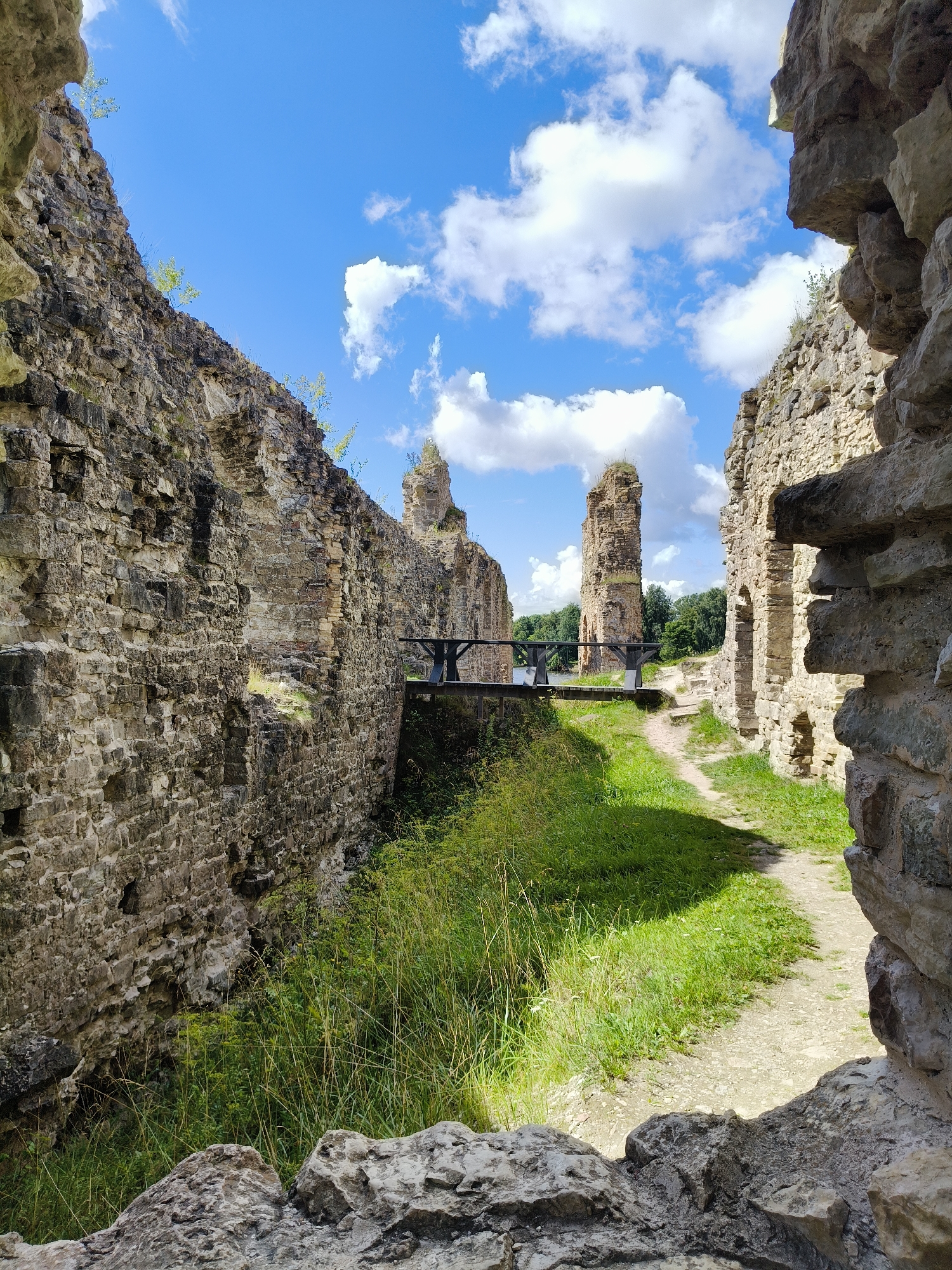 The image captures the ruins of Koknese Castle in Koknese, Latvia, under a bright blue sky with scattered white clouds. The ruins consist of tall, weathered stone walls, some of which are partially collapsed, creating a dramatic, somewhat overgrown landscape. A wooden bridge, supported by dark metal beams, spans across a lower area between two sections of the ruins, leading towards a path. The path winds through lush green grass and vegetation, suggesting a well-maintained historical site. The stone architecture is rough and textured, showing the effects of time and weather. The perspective is from within the ruins, looking out towards the bridge and the remnants of the castle structures beyond, framed by the rugged stone of the foreground. There are no people visible in the image. The scene is peaceful and evokes a sense of historical grandeur and decay.