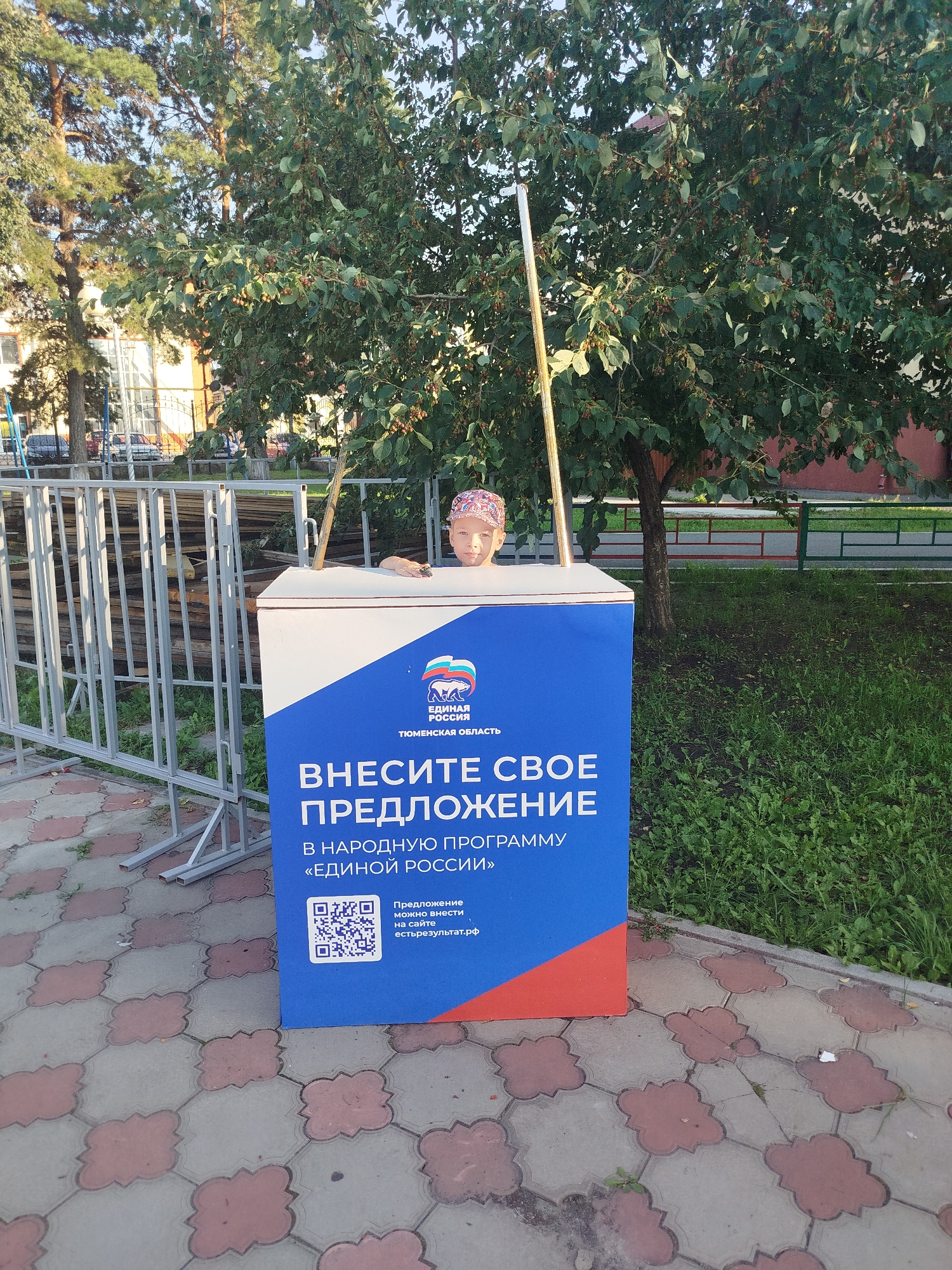 A young child wearing a patterned bandana on their head is peeking over the top of a white and blue donation box. The box has a large blue section with white lettering that reads "ВНЕСИТЕ СВОЕ ПРЕДЛОЖЕНИЕ" (Make your proposal). Below this, it says "В НАРОДНУЮ ПРОГРАММУ «ЕДИНОЙ РОССИИ»" (into the popular program of "United Russia"). To the left of the text, there is a QR code and the text "Предложение можно внести на сайте естьрезультат.рф" (Proposal can be made on the website estresultat.rf). A thin metal pole extends from the top of the box. The donation box is situated on a paved walkway with a repeating clover-like pattern. In the background, there are metal barriers, trees with green leaves, and a building. The scene appears to be outdoors, possibly in a public area of the city of Yalutorovsk, Russia, given the Cyrillic text on the box which references the Tyumen Oblast.