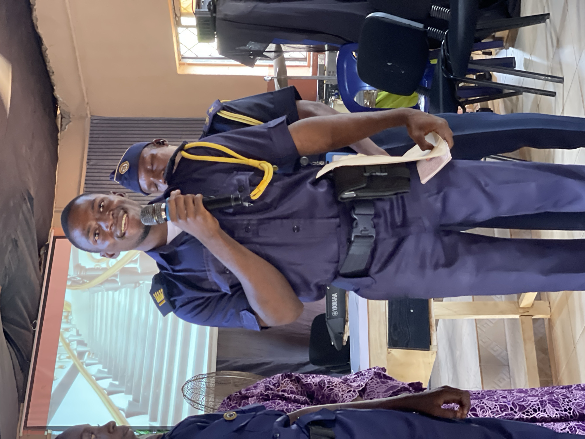 Akwanga, Nigeria, indoor scene: Two men, one holding a microphone, are in blue uniforms with yellow accents, potentially security or service personnel. The man with the microphone is speaking or presenting. Behind them is a projection screen displaying what appears to be a digitally rendered staircase. In the background, there are chairs, equipment cases, and a glimpse of another individual in uniform. A decorative cloth is draped in the foreground. The location suggests a public event or presentation space.