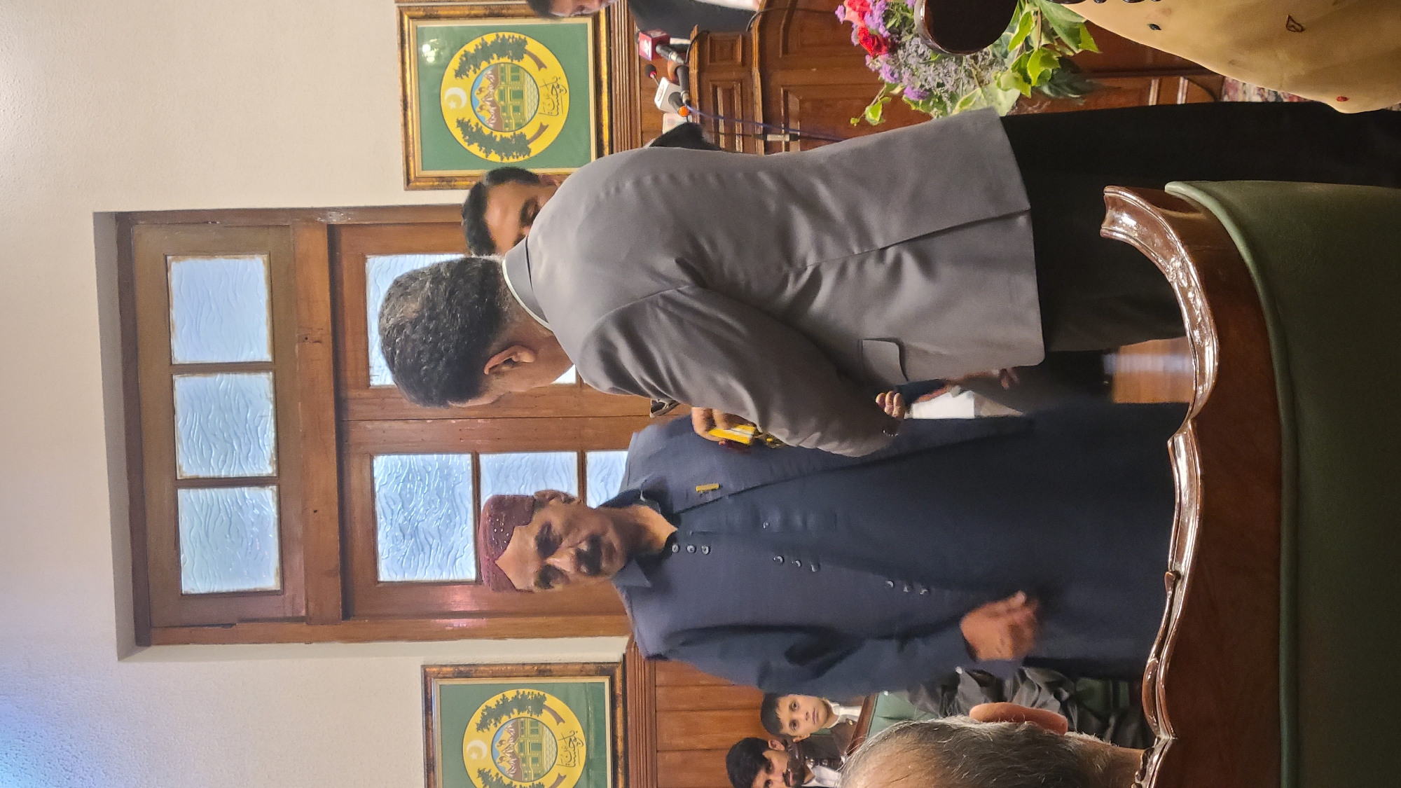 Captured indoors, the photograph displays a gathering of individuals. One man, clad in a dark navy traditional garment and a red hat, stands out. Another individual in a grey suit leans towards him. In the backdrop, a wooden window, framed wall paintings, and a gathering of spectators are visible. Given the green and yellow emblem on the wall art, this scene may be set in Quetta City Tehsil, Pakistan.