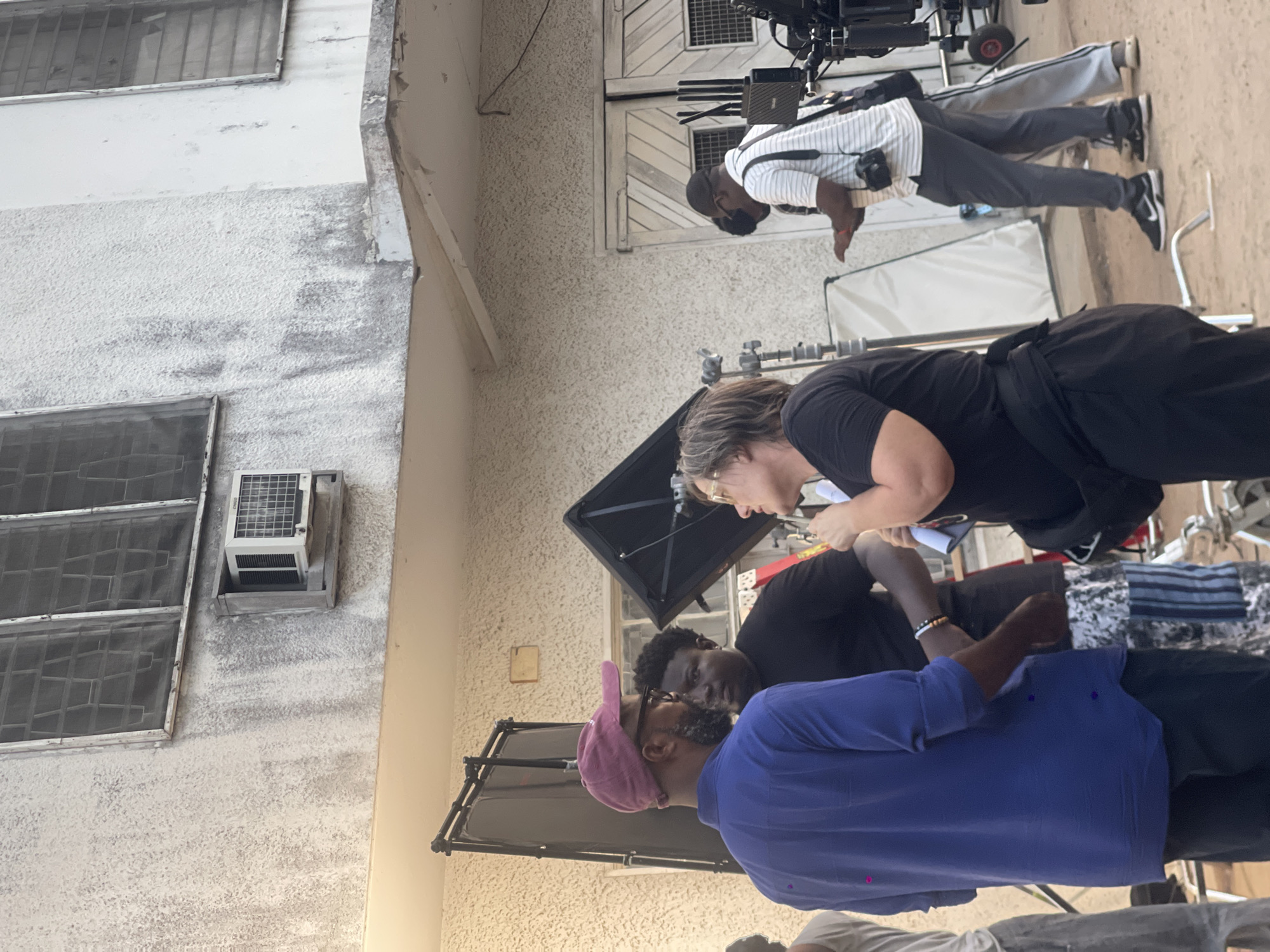 The image shows a film crew working on location in Unknown, Unknown. There are at least three people visible: a woman with light hair wearing a black shirt, a man with dark hair wearing a blue shirt and a pink hat, and a man wearing a black shirt. The crew is positioned around film equipment, including a large reflector screen and a camera on a dolly. The background features a white building with windows and an air conditioning unit. The setting appears to be outdoors during daylight.