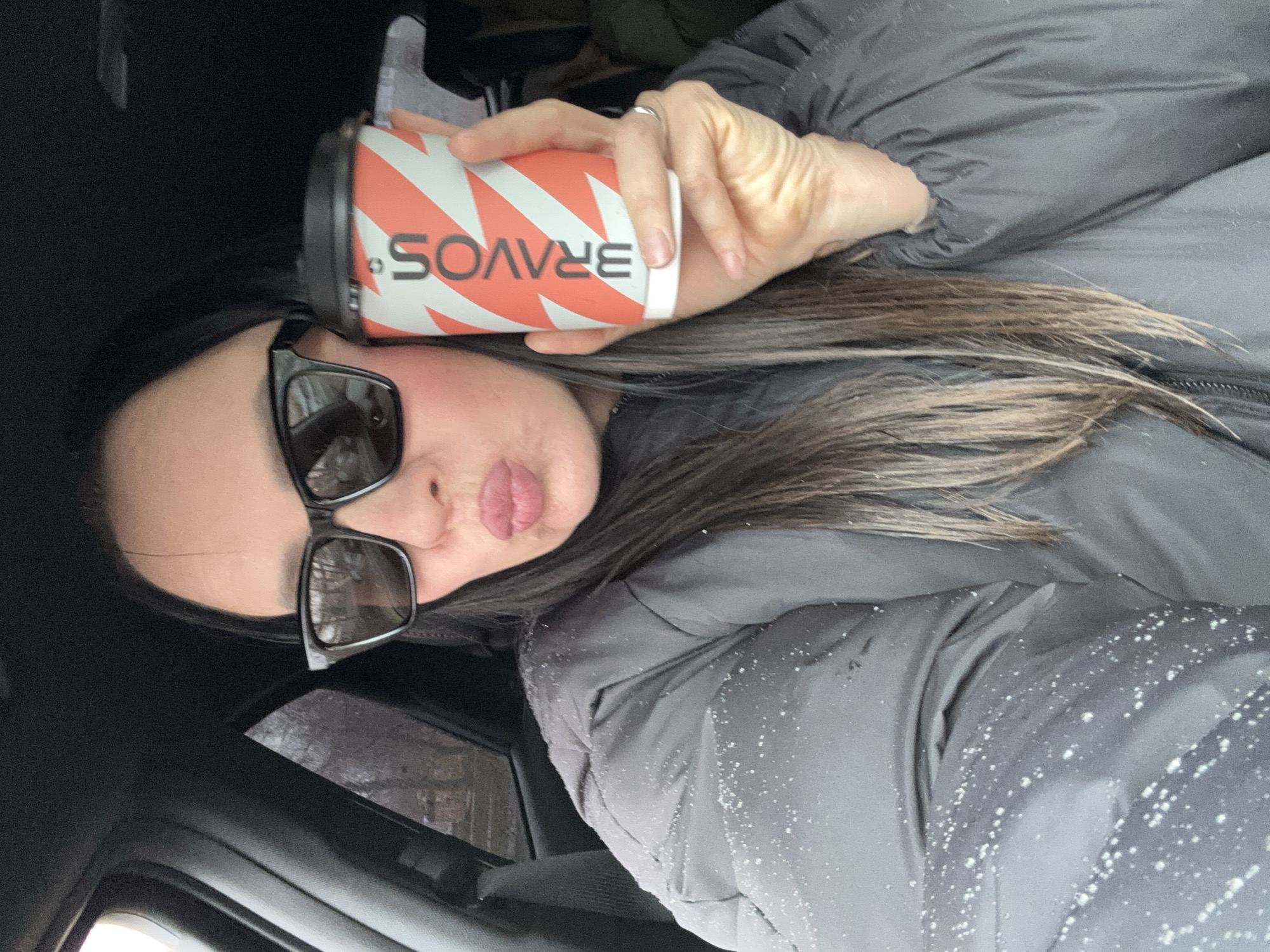 A woman is taking a selfie in a car, likely in Chaikovsky, Russia. She is wearing sunglasses and a gray puffer jacket with small white specks on it, possibly snow. She is holding a coffee cup with red and white angled stripes and the word "BRAVOS" printed on it. She is making a kissing face towards the camera. The interior of the car is visible, including the dashboard and door panel. A window is also visible, showing an out-of-focus background.