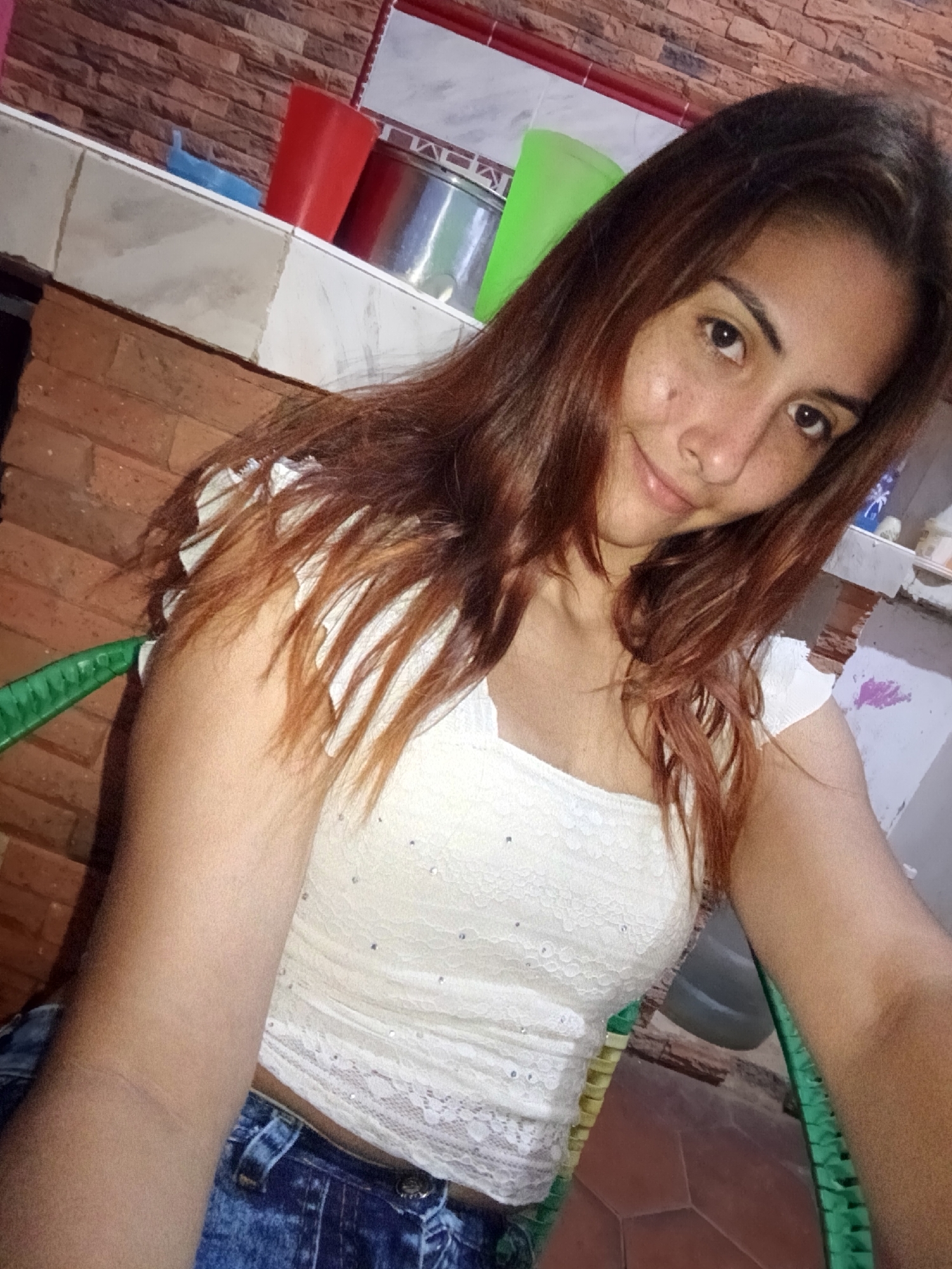 The image is a selfie taken by a young woman with long, auburn hair. She is wearing a white, lace-trimmed top with rhinestones. She has freckles on her face and is smiling at the camera. She is sitting in a green chair with a wicker back. In the background, there is a brick wall, a tiled surface, and some objects on a shelf, including red and green cups and metal containers. The floor appears to be tiled. The setting is likely indoors, possibly a kitchen or outdoor seating area, in Juan Griego, Venezuela.
