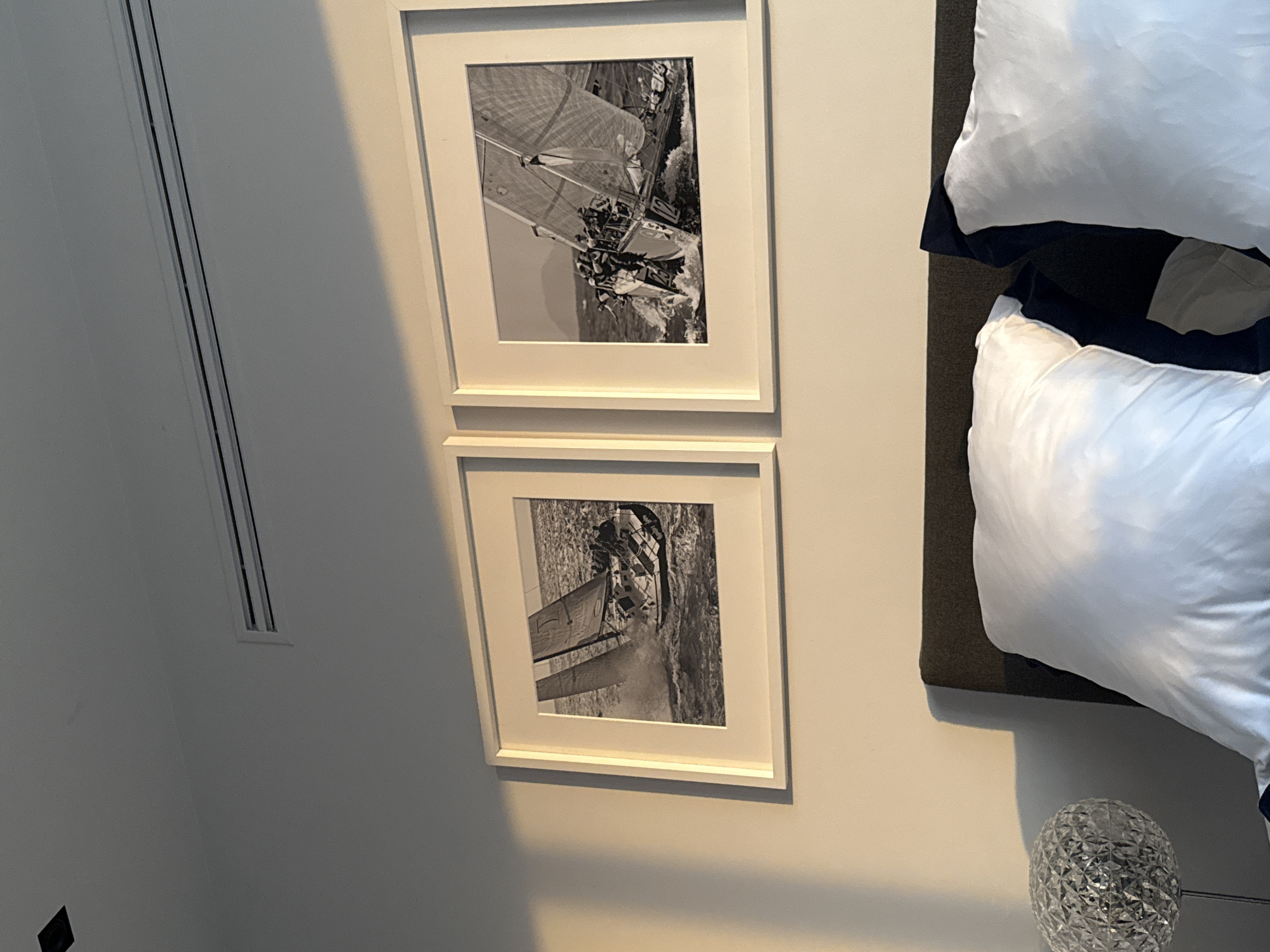 The image shows a room with two framed black and white photographs of sailboats hanging on a light-colored wall. Adjacent to the wall are a bed with white pillows and a bedside lamp with a textured glass base. The scene appears to be indoors, possibly in a bedroom or hotel room. The location is not identifiable as Mexico City, Mexico based on the provided image.