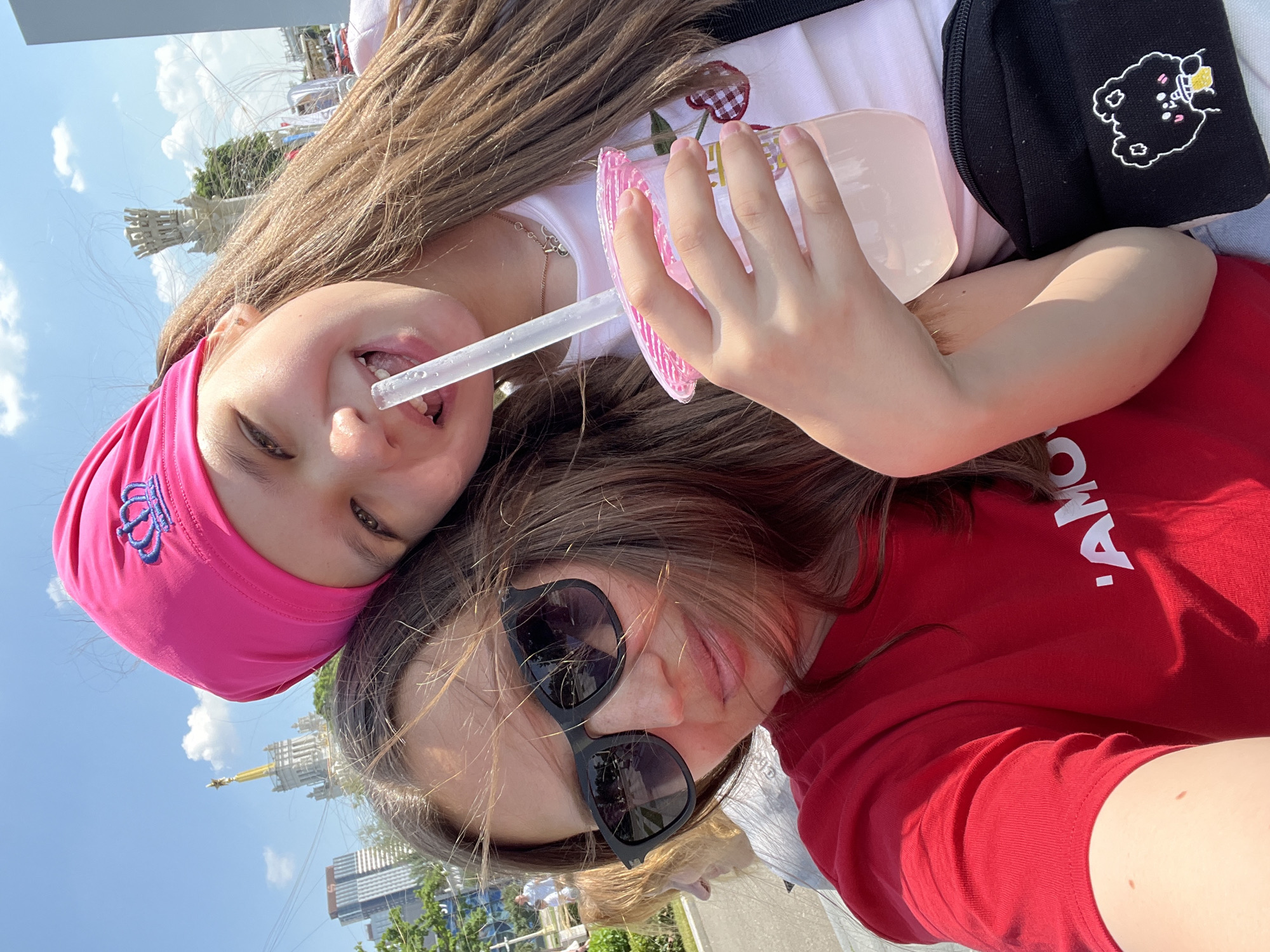 Two females are posing for a selfie outdoors on a sunny day. The female in the foreground is wearing a red t-shirt with white lettering and black sunglasses. Her hair is brown and windswept. The female behind her is wearing a pink bandana with a crown emblem and a white t-shirt. She is holding a pink cup with a straw in it and is drinking from it. The background appears to be a cityscape with tall buildings and a cloudy blue sky. The architecture suggests the location is likely in Moscow, Russia, possibly near a park or exhibition center with iconic structures like the Moscow State University or VDNH.