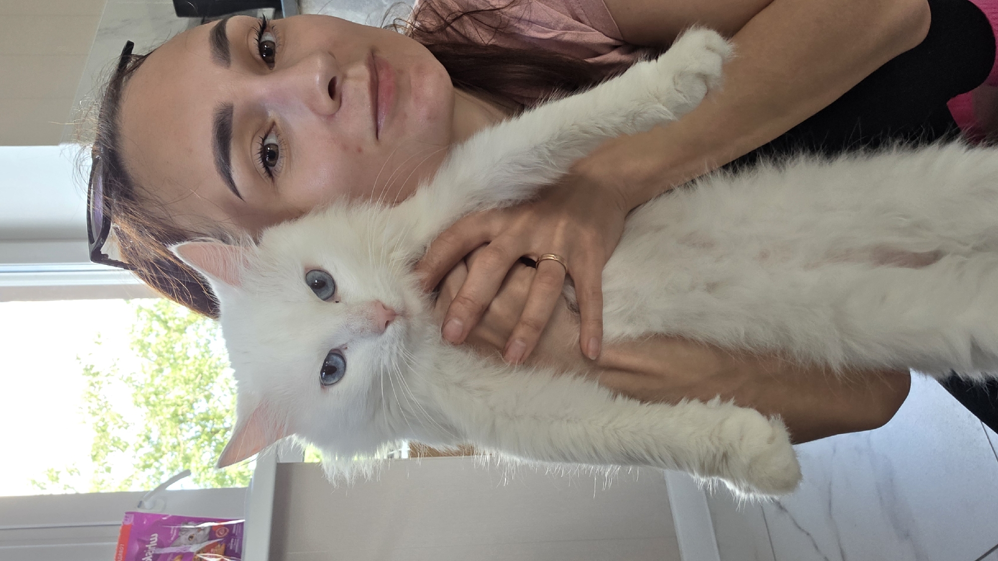 A woman holds a long-haired white cat with blue eyes. The woman has dark brown hair, worn up, and is wearing a pink shirt. Her face is partially visible as she looks towards the camera, with glasses perched on her head. The cat is held vertically in her arms, its body long and fluffy. The background shows a window with greenery visible outside, suggesting an indoor setting with natural light. The floor appears to be tiled. This scene is not located in Старошареево, Russia.