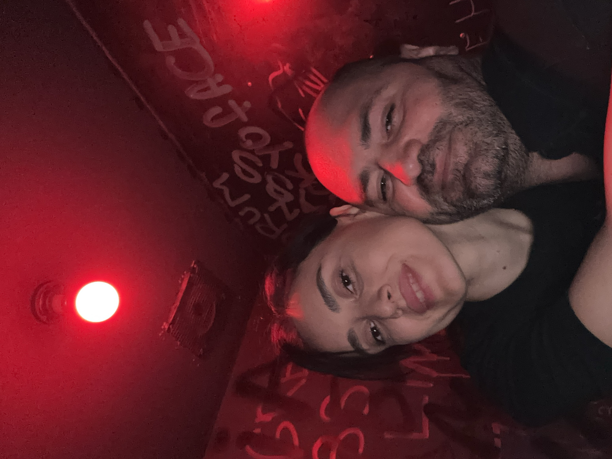 Two people, a man and a woman, are posed closely together under a red light. The man has a shaved head and a beard. Both appear to be wearing black shirts. The background appears to be a wall with graffiti in white lettering. A red light fixture is visible in the upper left of the frame. The location is unknown.