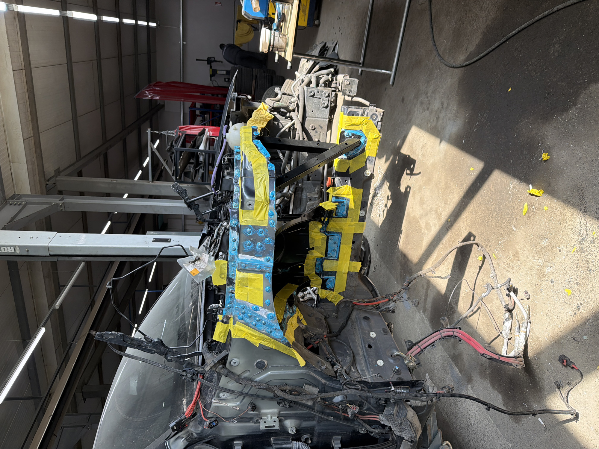 The image shows the front end of a car undergoing repair in a garage or workshop. The vehicle's front body panels are partially disassembled, revealing the internal structure. Various components, including wiring harnesses and possibly the frame, are exposed and covered in yellow tape, possibly for protection or marking purposes. The car is located indoors, with natural light entering from an unseen source. The background suggests a professional automotive repair environment, with machinery and tools visible. There are no people depicted in the immediate frame. There is no evidence in the photo connecting it to the city of Burgas, Bulgaria.