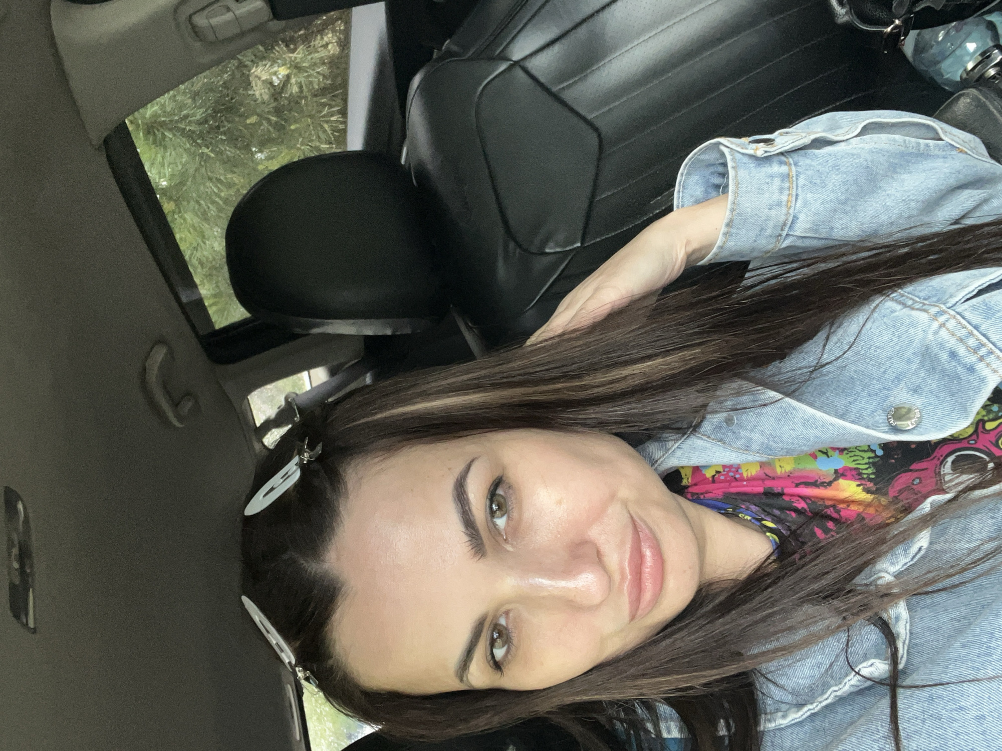 A woman is positioned inside a vehicle, possibly a car, as indicated by the black leather seats and car interior visible in the background. She has long brown hair, light skin, and is wearing a light blue denim jacket over a colorful shirt. The woman is looking directly at the camera with a slight smile. Two white hair clips are visible in her hair. Outside the vehicle, green trees can be seen through the window. The specific city and location are unknown.