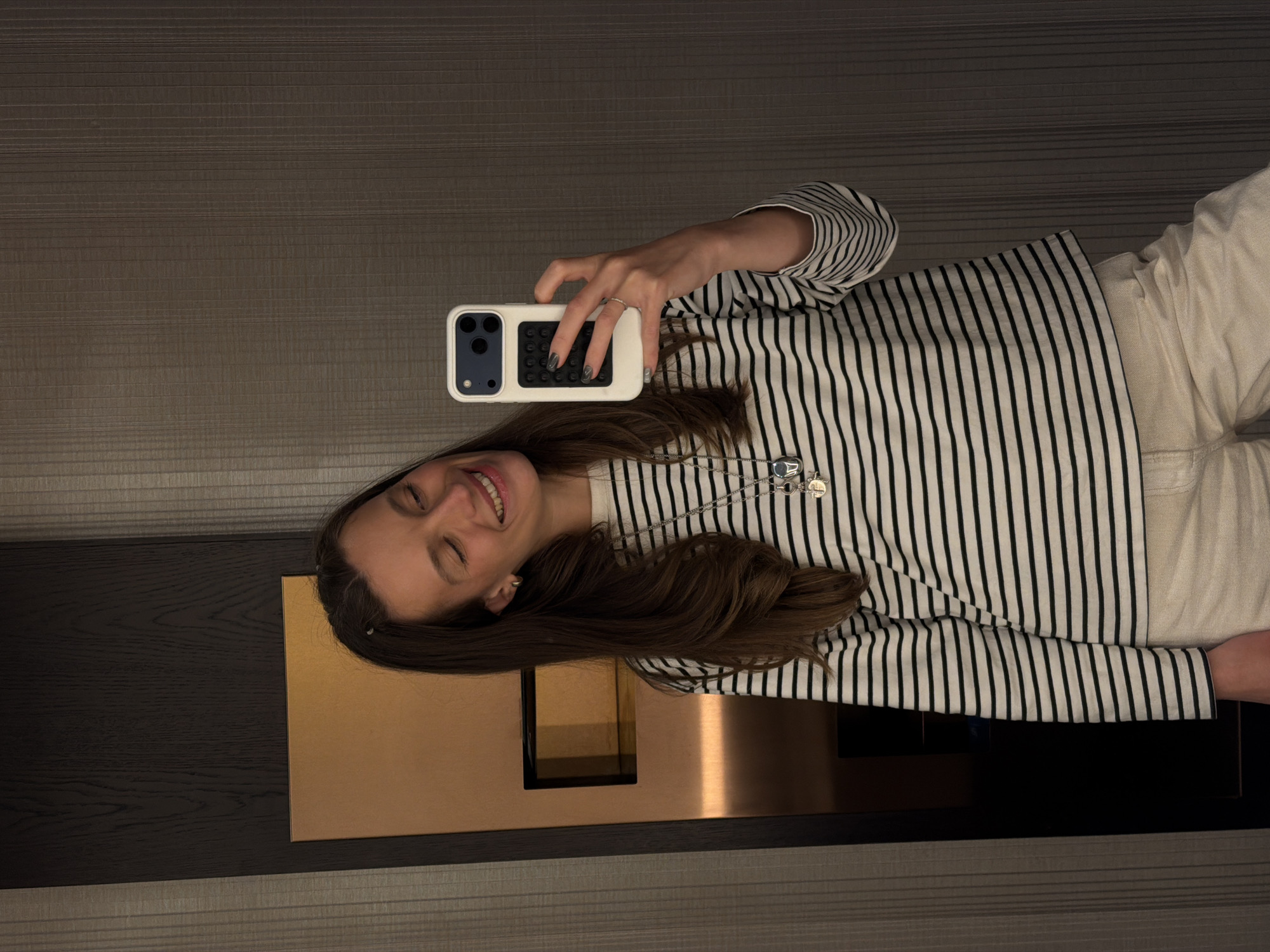 A woman is taking a selfie in a hotel room. She is wearing a black and white striped shirt, beige pants, and a silver necklace. She is holding a white iPhone with a black case. The room has a dark wood wall and a metallic gold accent panel. There is a wall with brown textured horizontal lines. There are no indicators of Budva, Montenegro present in the image.