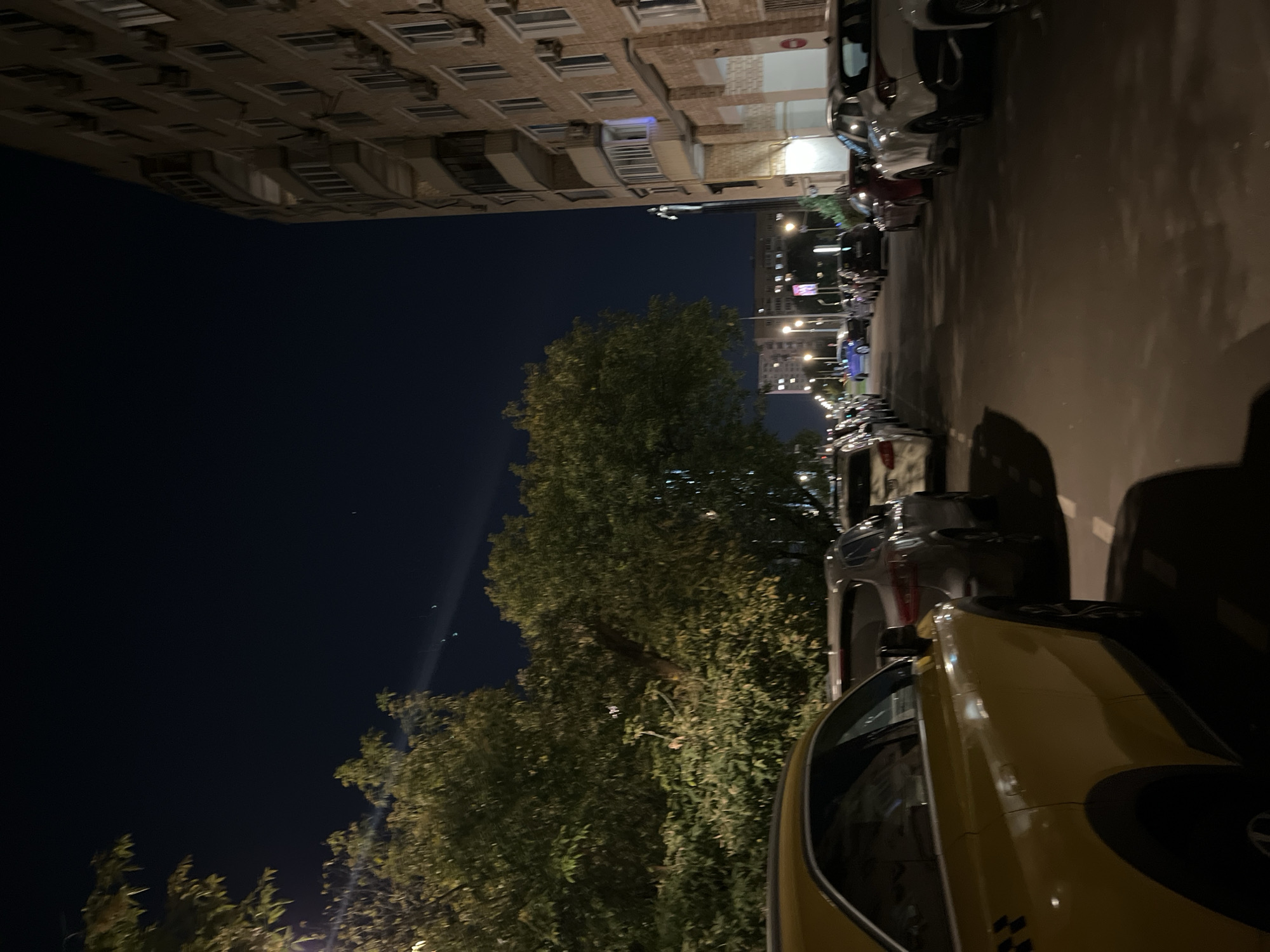 This is a night-time, eye-level, medium shot of a residential street in Moscow, Russia. On the right is a multi-story brick apartment building with numerous windows and air conditioning units. A line of parked cars runs along the street. In the foreground on the left is the rear of a yellow vehicle, possibly a taxi, with a black checkered pattern visible on its rear quarter panel. To the left of the street is a line of trees with dense foliage, some of which are illuminated by streetlights. The sky above is dark blue, with a few faint lights visible in the distance. The overall scene depicts a typical urban environment at night, with no people or overt activity present.