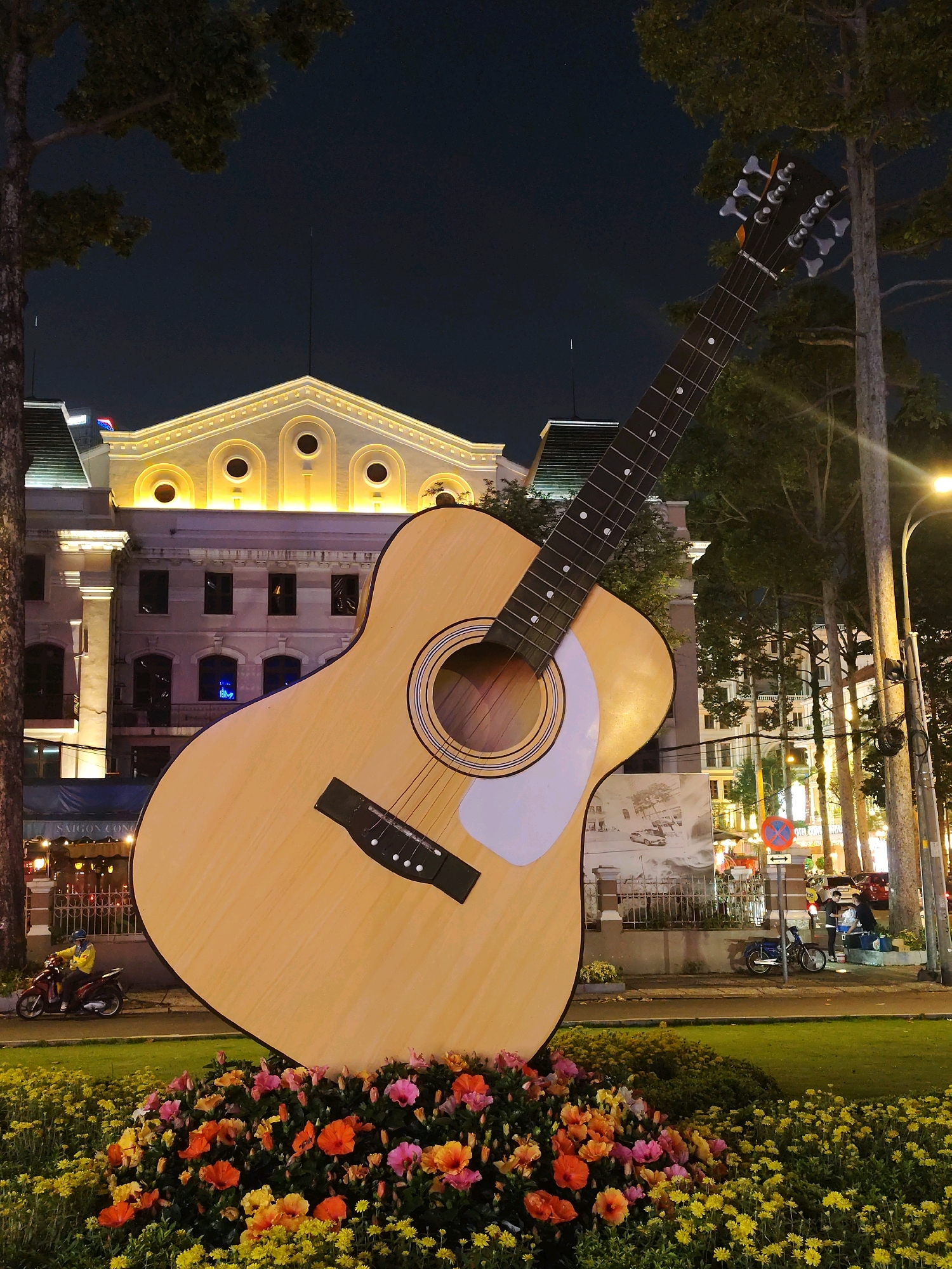 This is a low-angle, medium shot of a giant acoustic guitar sculpture placed on a bed of colorful flowers in front of a large, ornate building. The guitar is light brown with a white pickguard and black strings. The building, illuminated by warm yellow lights, appears to be a historical or cultural landmark, possibly a theater or opera house, with arched windows and decorative molding. The scene is set at night, with a dark sky visible above. Trees with dark foliage frame the left and right sides of the image. In the background, streetlights and traffic lights cast a glow. A person wearing a yellow jacket is riding a scooter past the flowers. Further back, a few people are gathered near a bicycle and what appears to be a food stall. The overall impression is of an artistic urban installation in Ho Chi Minh City, Vietnam, likely at night to highlight the illuminated building and sculpture. The location is identified as the Saigon Opera House (Nhà hát Thành phố Hồ Chí Minh).