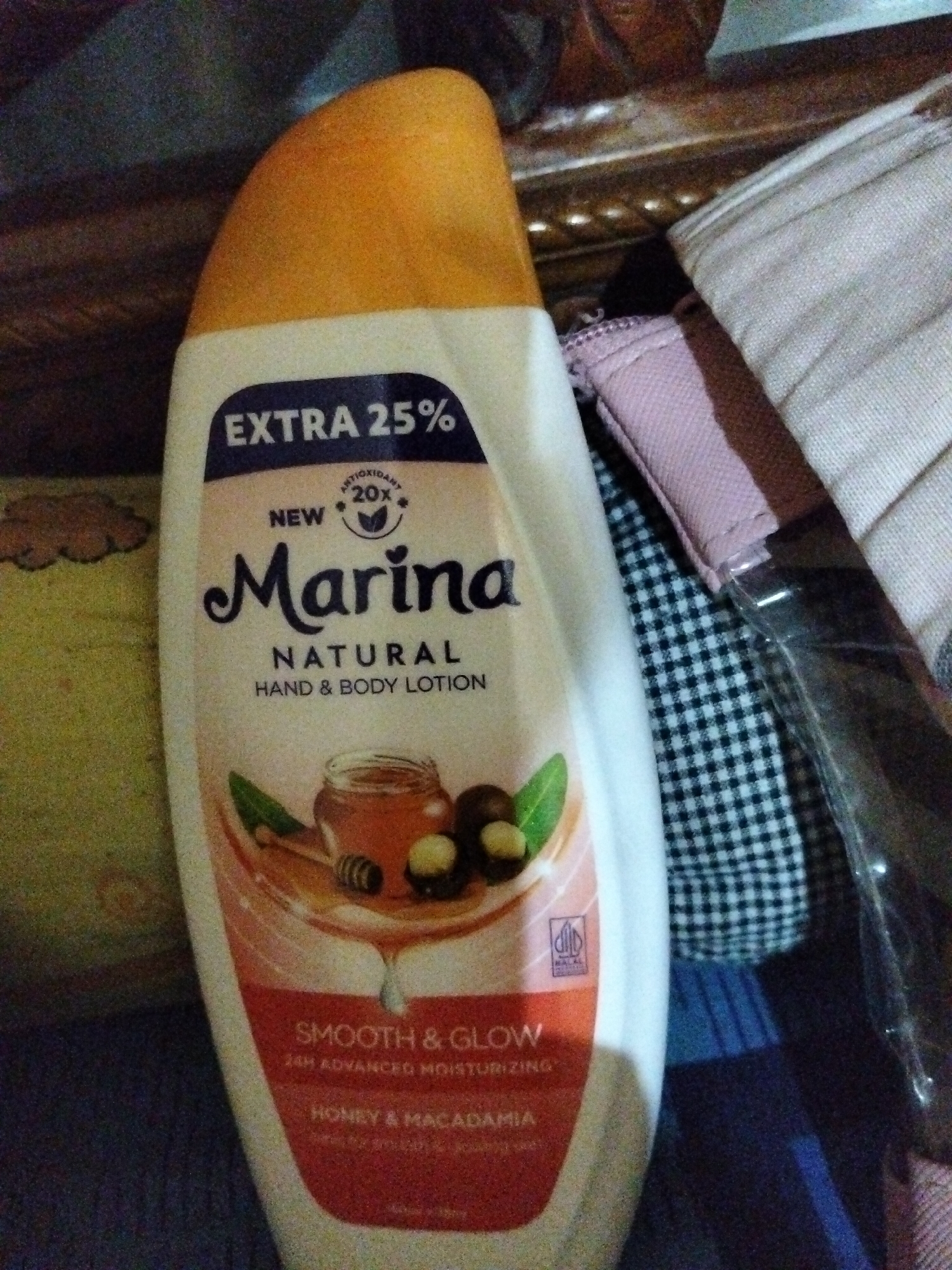 The image displays a bottle of Marina Natural Hand & Body Lotion with "Honey & Macadamia" scent, which appears to be an item within a household setting. The bottle is white with a bright orange cap and features a label with various text and graphics, including the brand name "Marina" in a stylized font. There are also graphics of honey, macadamia nuts, and leaves. The bottle is positioned on a surface, possibly a table or shelf, and is partially obscured by other objects such as fabric items with checkered patterns and solid colors. The lighting suggests an indoor environment. The image does not provide any information about the city of Payagaboh, Indonesia, nor does it depict any people, actions, or interactions.