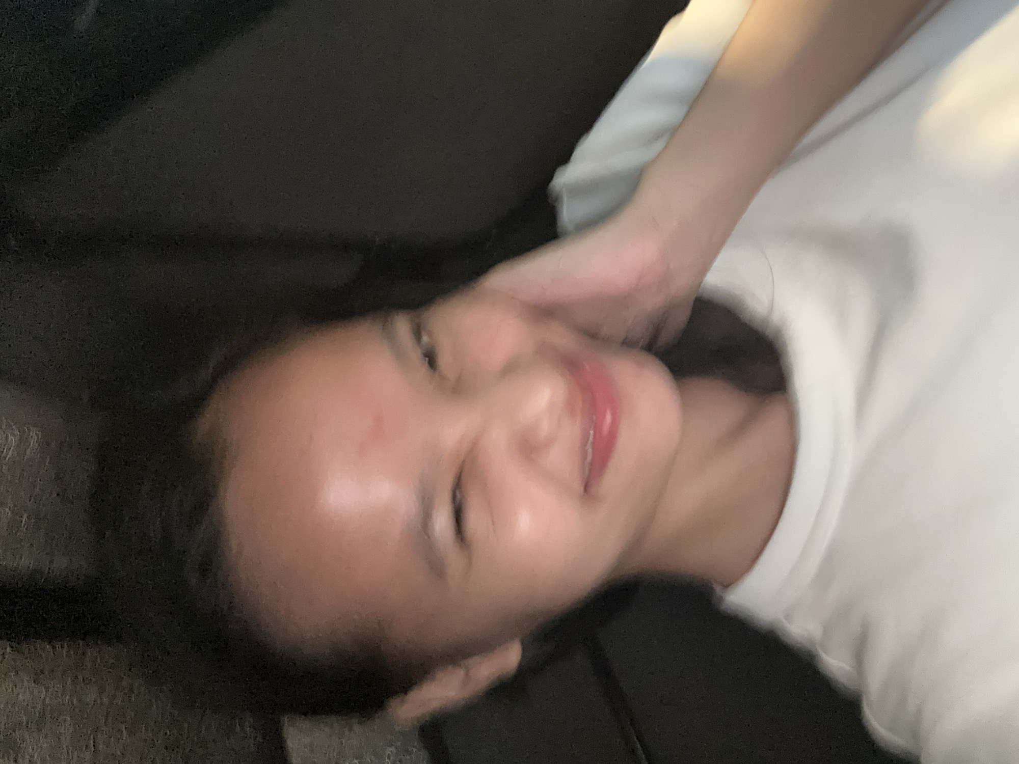 The image shows a close-up, selfie-style shot of a young woman with her right hand touching her cheek. She is wearing a white t-shirt. Her face is partially obscured by her hand and she is winking, with her mouth open in a smile. The background appears to be the interior of a vehicle, with dark upholstery visible. There is no identifiable information or objects in the image that would place its location in Davao City, Philippines.