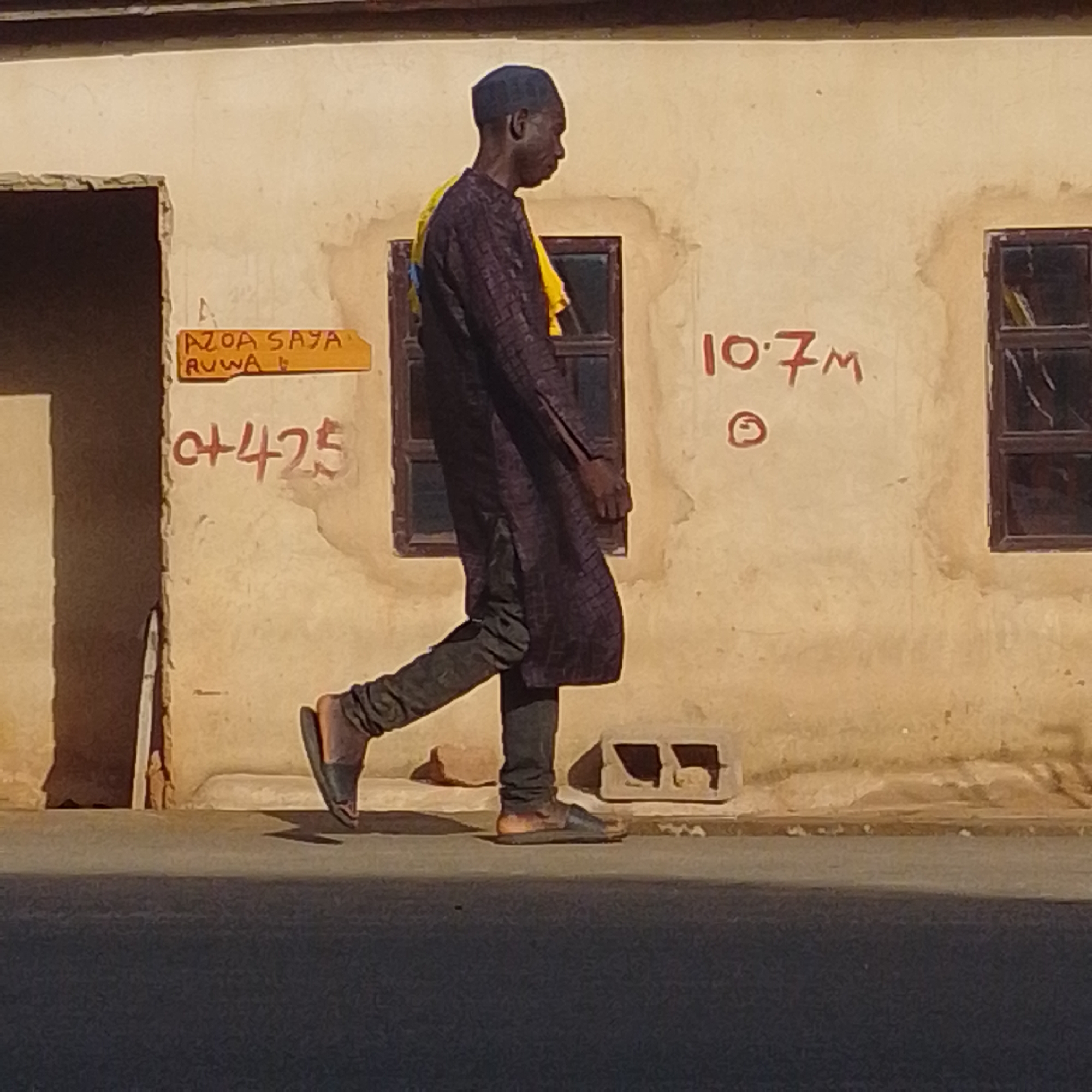 The image captures a side view of a man walking in front of a building with a stucco-like wall. The man is wearing a dark, patterned robe over dark trousers and dark sandals. He has a dark skullcap on his head. He is walking to the right of the frame, and his body is angled away from the viewer.
The building wall is light-colored and appears weathered, with some staining and peeling paint. There are several markings on the wall. To the left, a yellow sign is mounted, with text that appears to be "AZOA SAYA RUWA". Below this, red spray paint forms the characters "0+425". To the right of the man, more red spray paint reads "10.7M", and below that, a simple circular symbol. A dark-colored window frame is visible to the right of the man, with what appears to be glass that is broken or reflective. Near the bottom of the wall, two rectangular openings, possibly from cinder blocks, are visible.
The man is walking on a paved surface, which is in shadow, suggesting it is either early morning or late afternoon. The lighting indicates a sunny day. The scene appears to be in an outdoor, urban or semi-urban environment. The overall impression is one of everyday life in a warm climate, possibly in Gombe, Nigeria, as suggested by the location context. No specific emotions are clearly discernible from the man's profile, but his posture suggests purposeful movement.