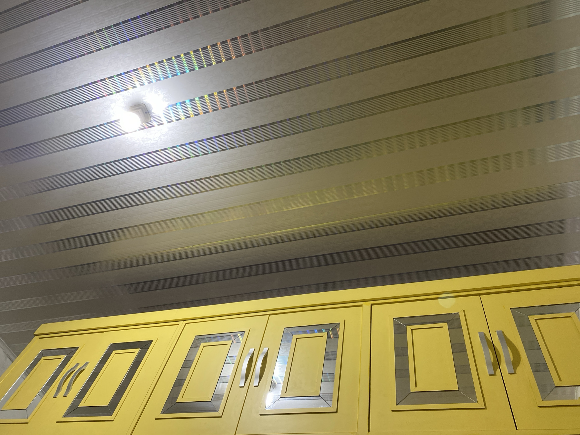 The image shows a low-angle shot of the ceiling and the top of yellow cabinets in an indoor environment. The ceiling is made of parallel, horizontal panels with a subtle, textured pattern and alternating bands of reflective material that create a rainbow effect when light hits them. A single, bright light fixture is visible on the ceiling, illuminating the area. Below the ceiling are the tops of yellow cabinets with decorative rectangular insets framed by a dark border. Some of these insets appear to have reflective or mirrored surfaces, catching the light from the ceiling fixture. Two silver-colored cabinet handles are visible on the front of the cabinets. The overall impression is that of a modern kitchen or storage area.
