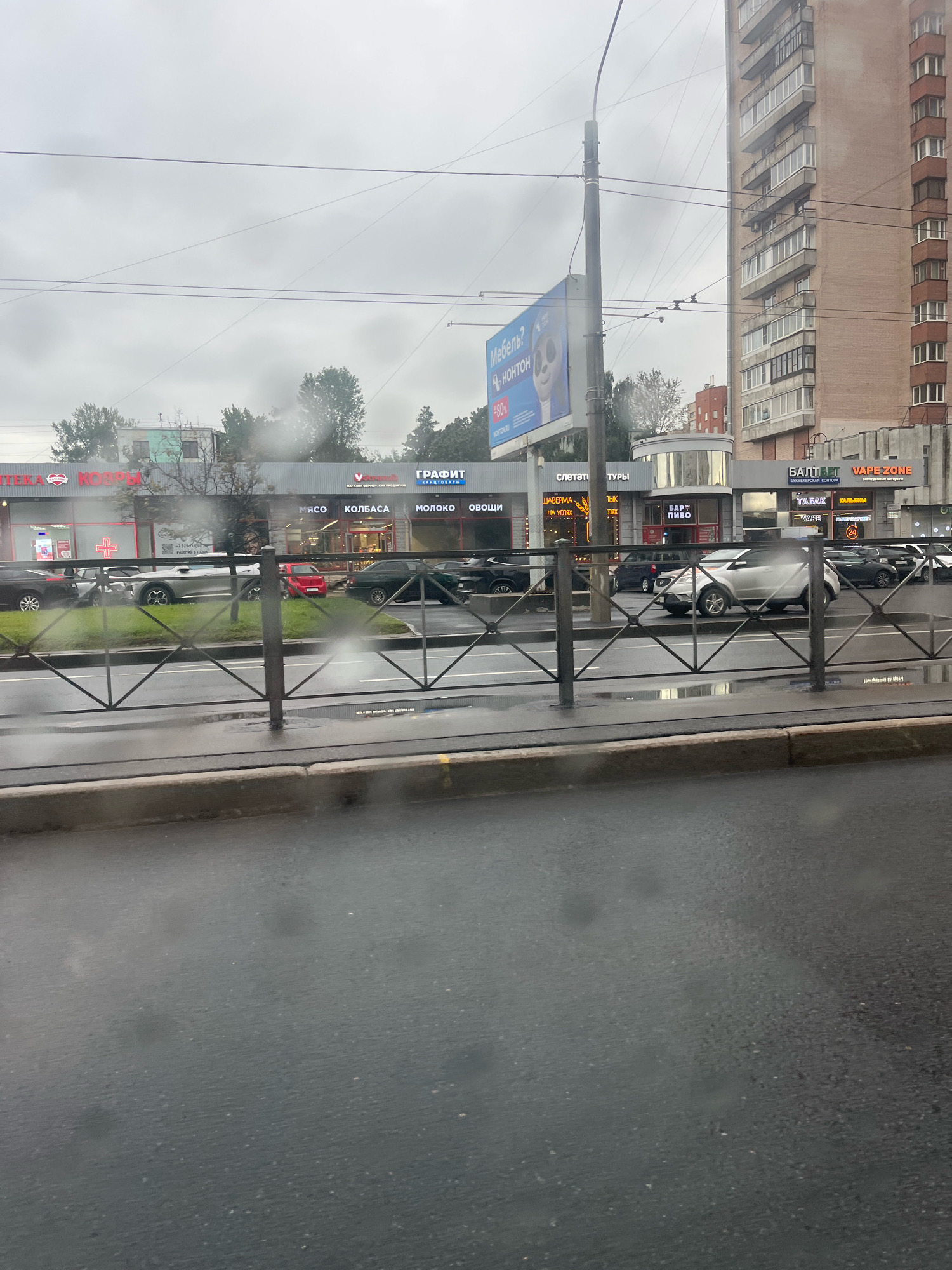 The image shows a cloudy day in Saint Petersburg, Russia. In the foreground, a wet road surface is visible with raindrops on a glass surface, likely from inside a vehicle. A metal fence separates the road from the sidewalk. Behind the fence, there is a line of shops and businesses. Visible signs include "АПТЕКА" (Pharmacy), "МЯСО" (Meat), "КОЛБАСА" (Sausage), "МОЛОКО" (Milk), "ОВОЩИ" (Vegetables), "ГРАФИТ" (Graphite), "БАЛТВЕТ" (BALTVET), and "VAPE ZONE". There are several cars parked and driving on the road. A tall apartment building is visible in the background, characteristic of the city's architecture. The overall atmosphere is gloomy and wet, suggesting it is raining or has recently rained.