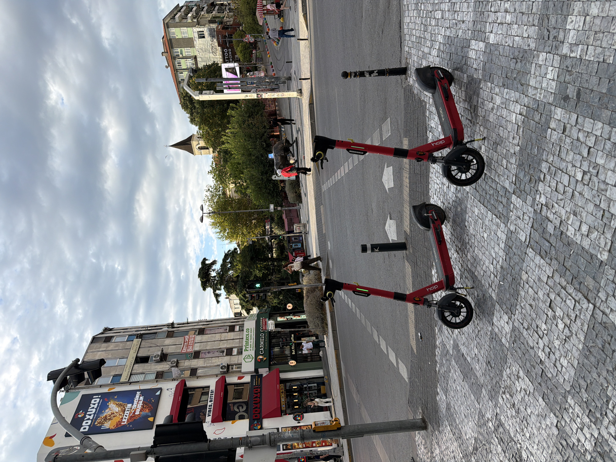In Kadıköy, Turkey, two red electric scooters are parked on a cobblestone crosswalk on a street with a cloudy sky overhead. In the background, a multi-story building features signage for "DOYUXO!", "VIBES, BITES!", "Printes.co", and "CARMELO COFFEE". Further down the street, people are walking, and a statue of a bull is visible. Another building with a church steeple is visible in the distance. Traffic lights are also present, with one showing green. The overall scene depicts a busy urban environment with elements of commerce and public spaces.