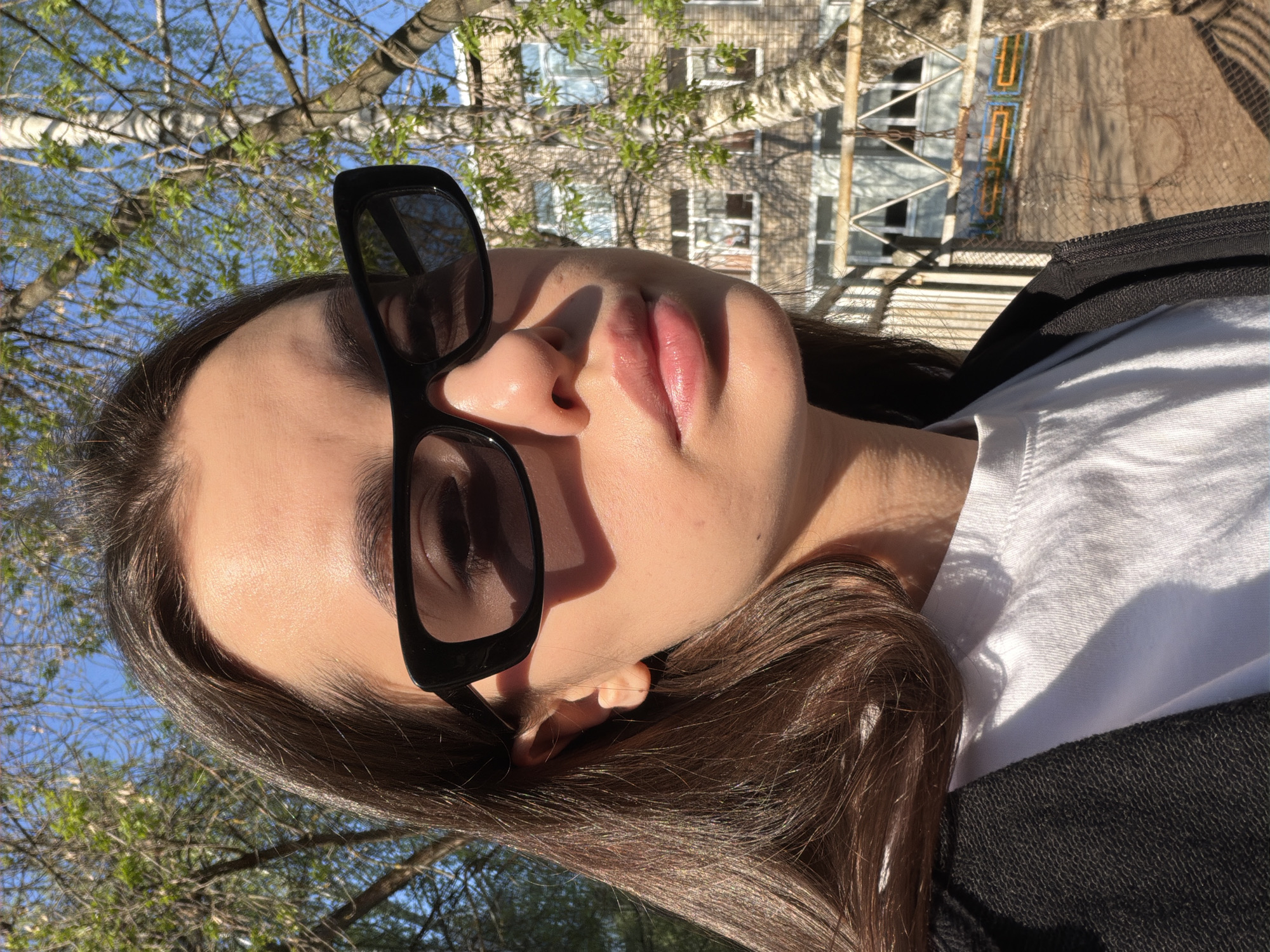 A person is posing for a selfie outdoors on a sunny day. The person has fair skin, dark hair, and is wearing dark sunglasses. The person is wearing a white shirt and a black jacket. In the background, there is a building and trees. The building is located in the city of Chaikovsky, Russia.