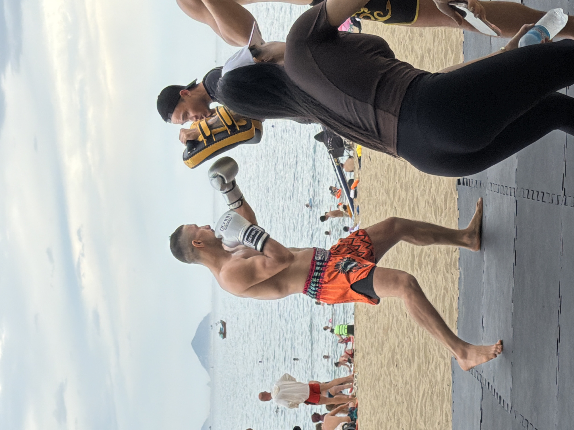 A boxing training session is taking place on a mat on the sandy beach in front of the ocean. Two men are actively engaged in the training: one is wearing boxing gloves and orange Muay Thai shorts, and is in a defensive stance, while the other, wearing a black cap, is holding a yellow and black punch pad. In the background, there are several other people on the beach, some are swimming or sunbathing. The ocean is visible with a boat in the distance, and a hill can be seen on the horizon. The sky is overcast. The overall scene suggests an outdoor fitness activity in Nha Trang, Vietnam.