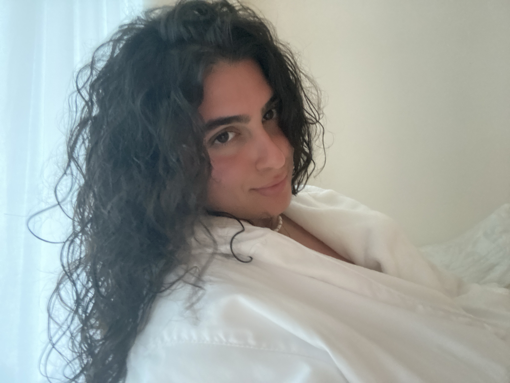 A person with long, dark, curly hair is looking at the camera with a slight smile. They are wearing a white shirt and a pearl necklace. The background is blurry but appears to be a white wall and a window with a sheer white curtain. The image does not provide sufficient information to determine the location in El Caracol, Mexico.
