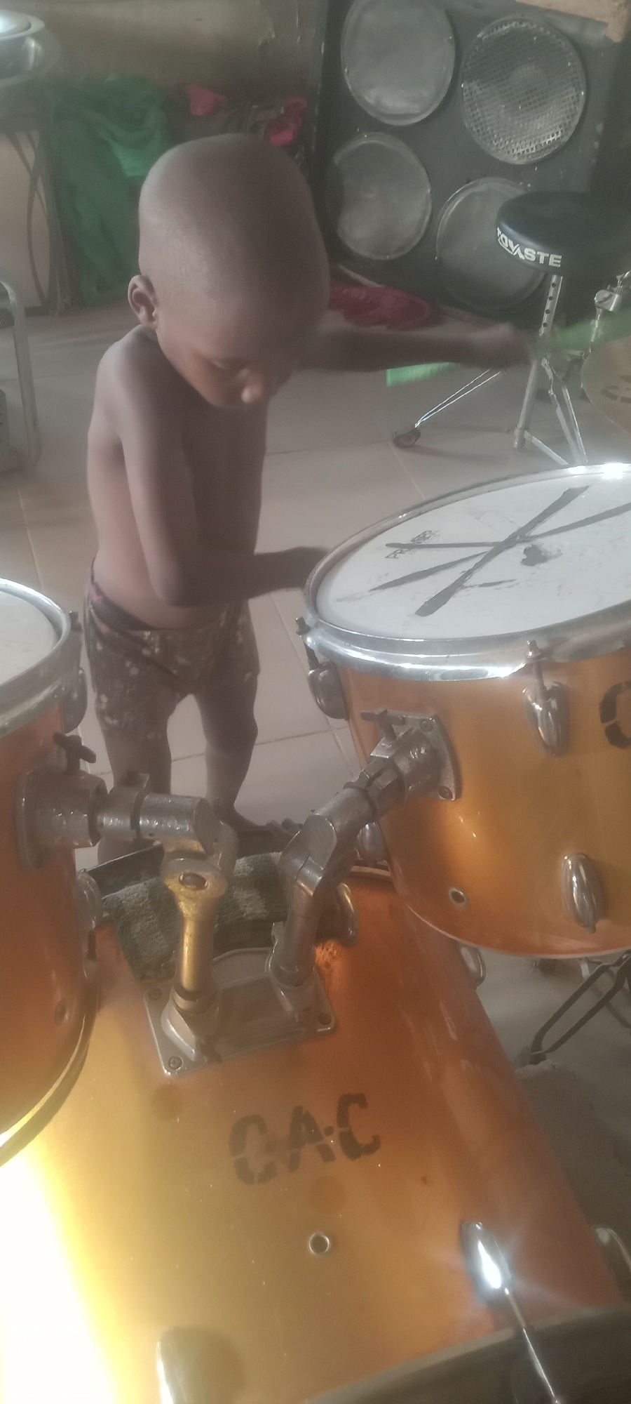 A child stands in a room and appears to be playing a drum set. The child is wearing patterned shorts and has a shaved head. The drums are orange-gold in color with the letters "CAC" visible on one of the drums. In the background, there is a speaker cabinet with multiple round speakers and a drum stool. The location is Bende, Nigeria.