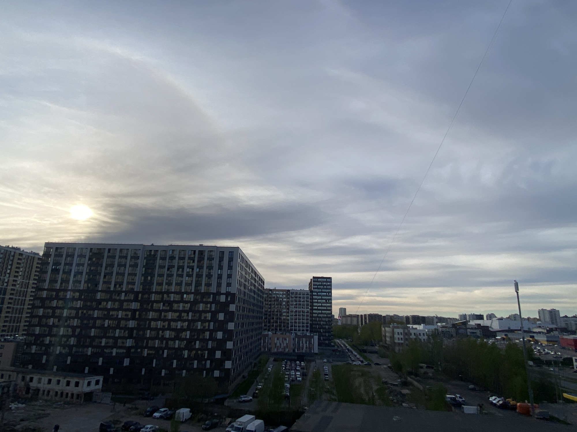 The image depicts an urban landscape, likely in Saint Petersburg, Russia, based on the architectural style and city layout. The scene is dominated by modern apartment buildings, with a large, multi-story residential complex taking up a significant portion of the left side. The building has a dark facade with numerous windows. Adjacent to the buildings are parking areas filled with cars and green spaces with trees. A few older, dilapidated structures are visible near the foreground, suggesting redevelopment or contrasting architectural styles. In the background, the skyline features a mix of other high-rise buildings and urban infrastructure, including a communication tower. The sky is overcast with thick clouds, and the sun is partially obscured, creating diffused lighting. A thin wire stretches across the sky, possibly a utility line. There is no visible evidence of people in the image.
