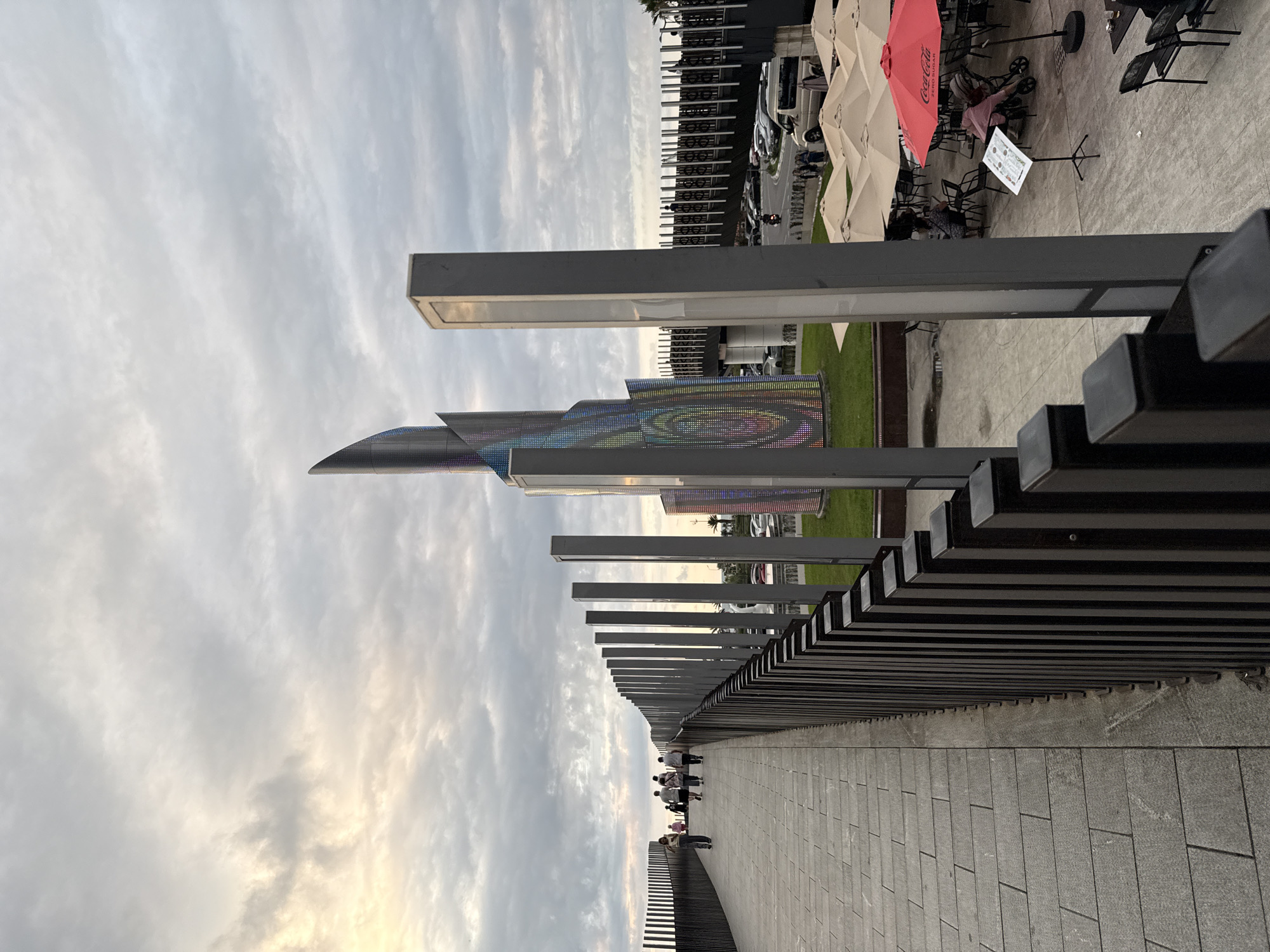 An outdoor perspective shot shows a pedestrian walkway on the left, leading towards a modern sculpture. A dark metal railing borders the walkway on the right. Several people are walking on the walkway, heading towards the sculpture. The sculpture is tall and cylindrical with a pointed top, featuring a colorful, pixelated pattern. In the background, there are cars parked on a road, some people gathered at tables under umbrellas, and a red umbrella with a Coca-Cola logo. The sky is cloudy with hints of sunset light.