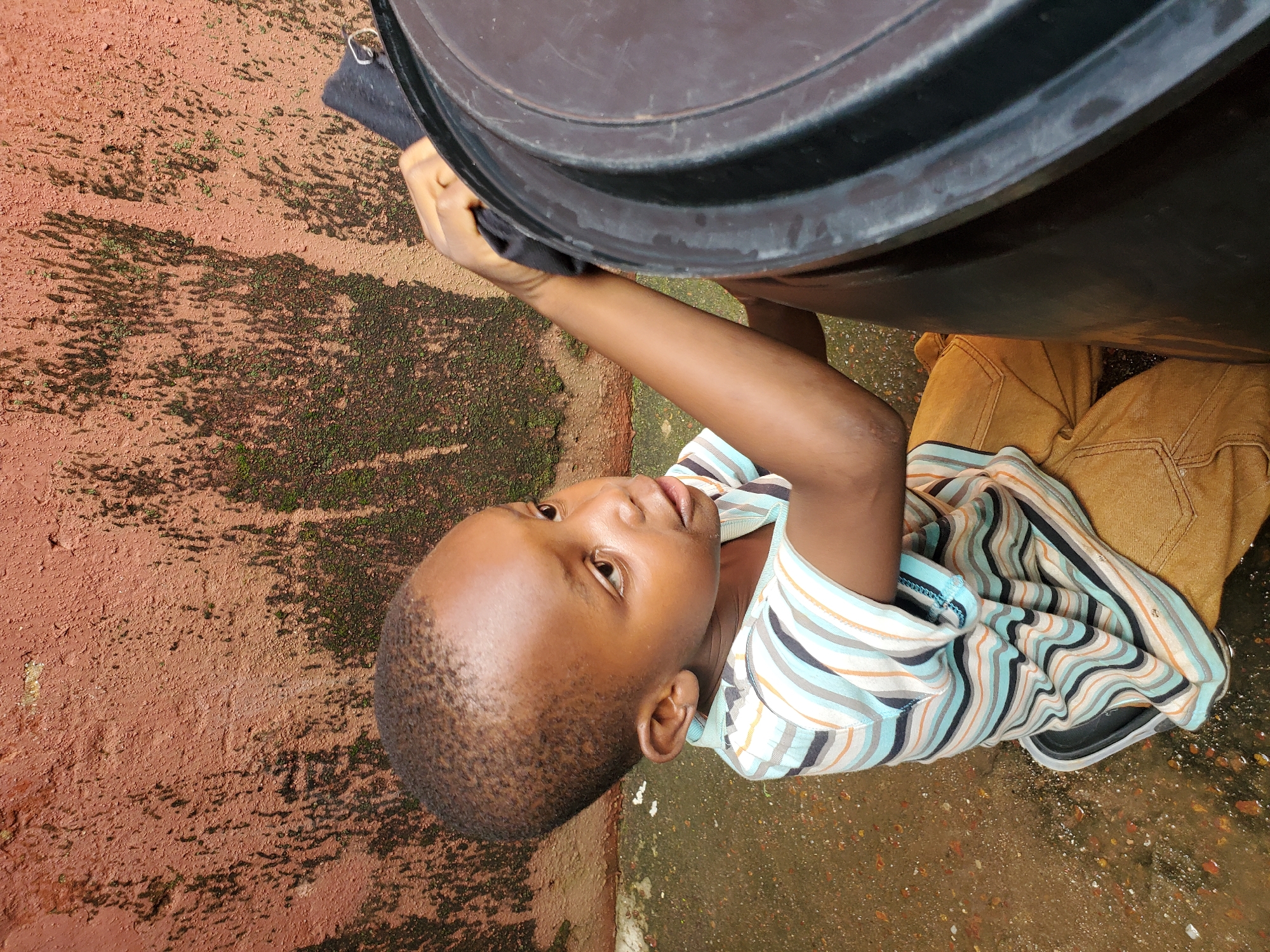 A young boy, likely of African descent, is captured in a close-up shot, oriented sideways. He is wearing a horizontally striped t-shirt with bands of white, blue, and orange, and brown trousers. The boy is leaning against a large, dark, cylindrical object, possibly a water tank, with his right arm extended and hand gripping the edge of the object. His gaze is directed upwards and slightly to the left, with a curious or thoughtful expression on his face.
The ground beneath him appears to be a reddish-brown, possibly dirt or clay, with patches of green moss or algae, suggesting a damp or humid environment. The texture of the ground is rough and uneven. Shadows are cast on the ground, indicating it is daytime, with the sun likely at a moderate angle. The overall lighting suggests an outdoor setting, possibly in a backyard or open area.
The image is shot from a high angle, looking down at the boy and the object he is interacting with. The context of Jos, Nigeria, suggests a warm climate. There are no visible text elements in the image. The activity depicted is simple and everyday, with the boy engaging with his surroundings. The focus on the boy's face and his interaction with the object creates a sense of intimate observation.