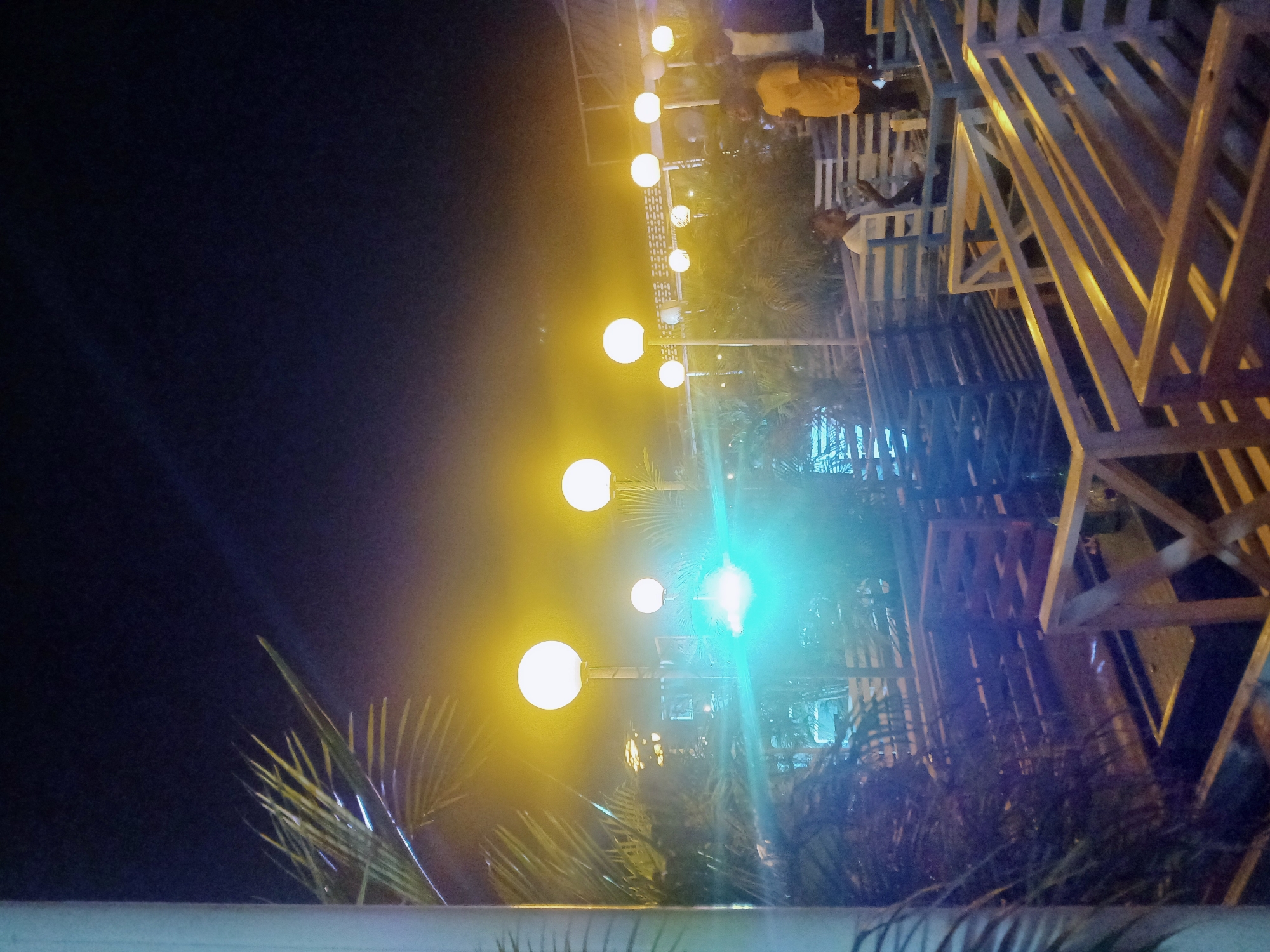 The image depicts an outdoor scene at night in Katambe, Nigeria. A string of spherical lights is visible, with one light emitting a bright blue hue. There is decorative geometric wooden construction on the right and a palm tree in the foreground. In the background, people are sitting at tables. The overall lighting is dim, with darker areas dominating the upper left portion of the image.