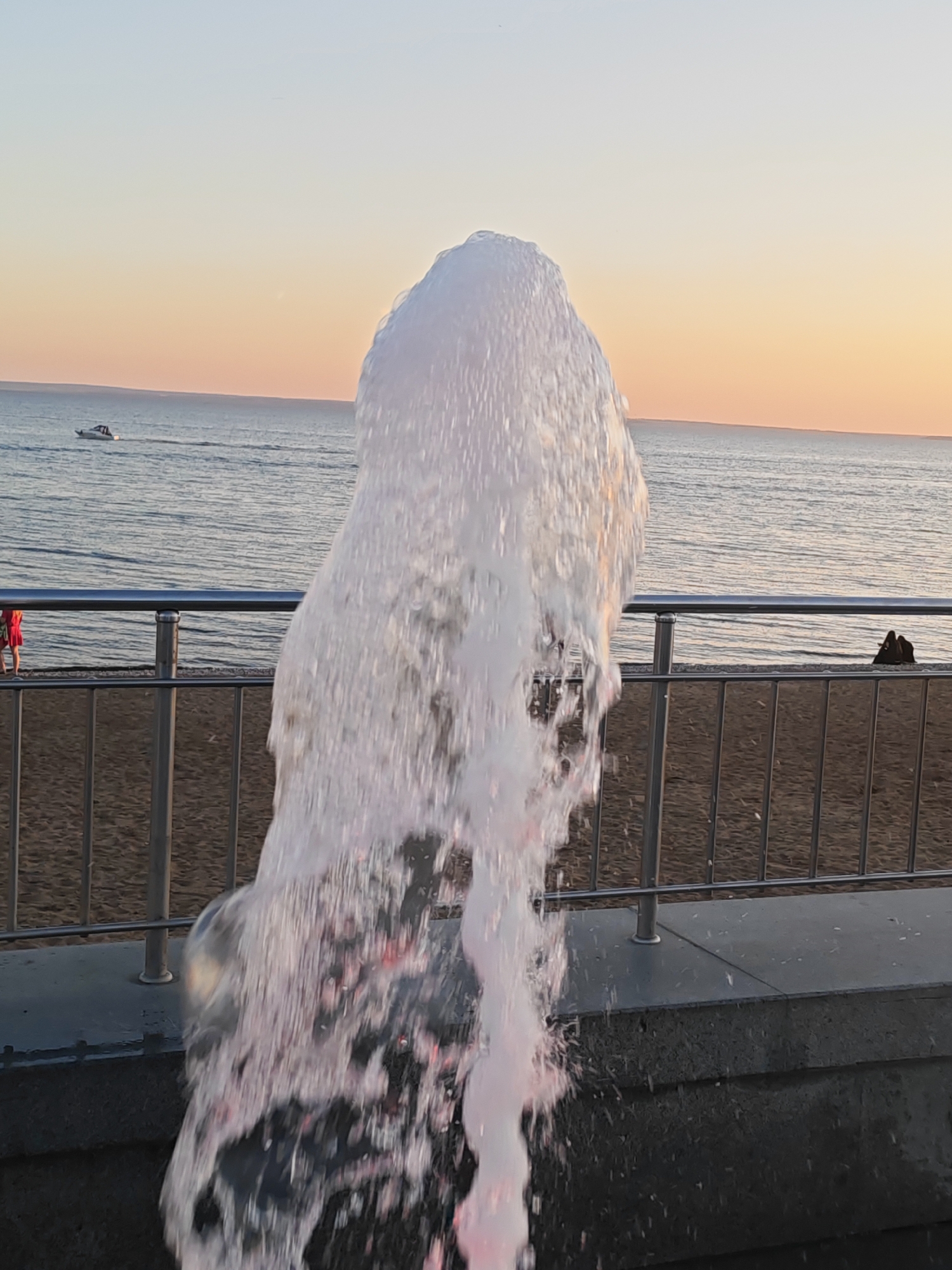 The image shows a fountain of water in the foreground, with a beach and the sea in the background. The sky is a gradient of orange and blue, suggesting it is either sunrise or sunset. There is a railing in the middle ground, separating the fountain area from the beach. On the beach, two figures are sitting, facing the sea. In the distance, a small boat is visible on the water. The scene appears to be at a waterfront location in Tolyatti, Russia.