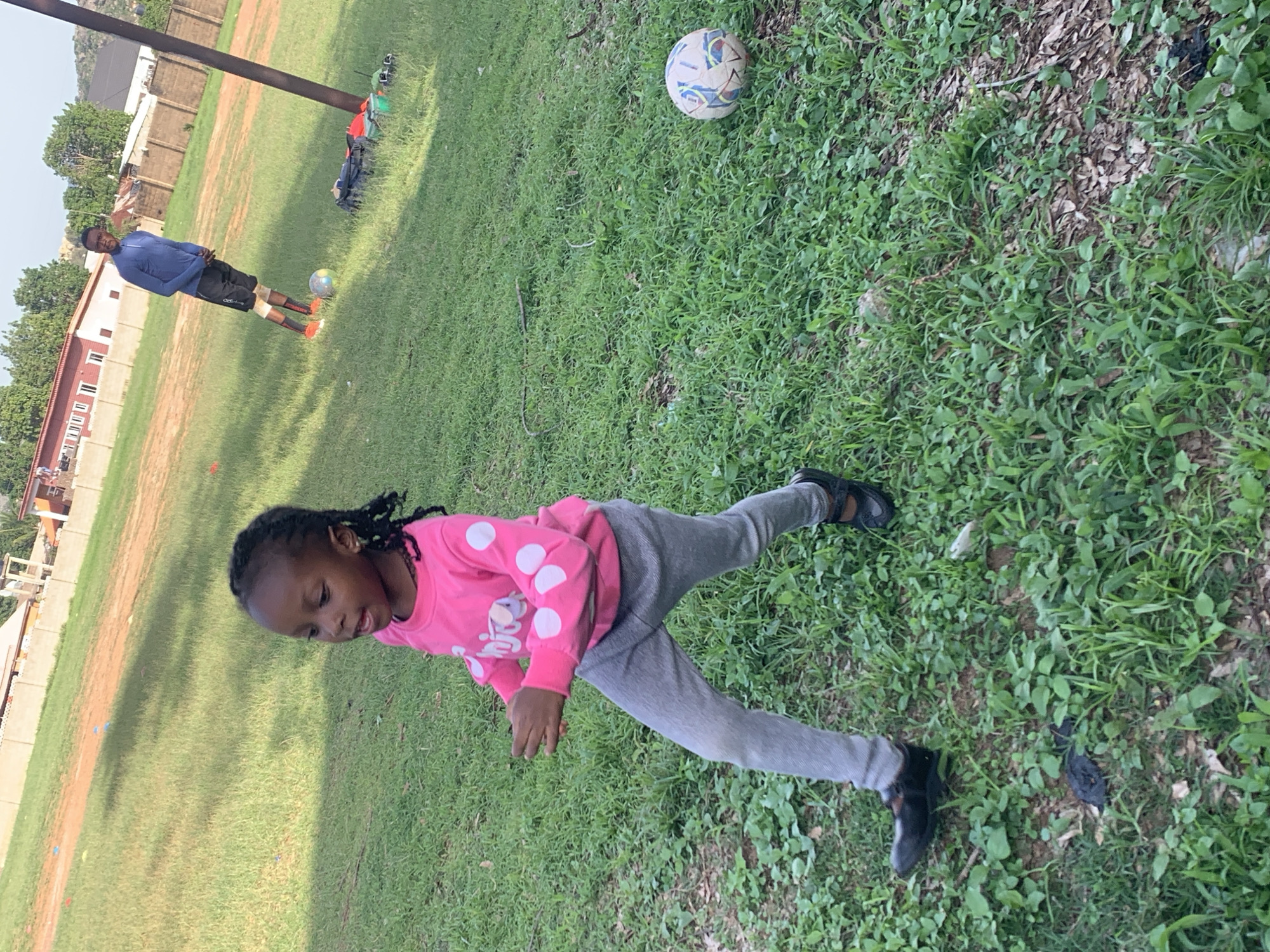 In Jos, Nigeria, on a grassy field with some dirt patches, a young Black girl with braided hair runs towards the left of the frame. She wears a bright pink long-sleeved shirt with white polka dots and gray leggings, and black shoes. Her mouth is open as if she is speaking or about to speak. In the background, a Black man in a blue long-sleeved shirt and black shorts stands looking towards the left. Next to him is a soccer ball. A tall, dark pole stands to the right of the man. The environment appears to be an outdoor recreational area, possibly a sports field, with some buildings and a fence visible in the background, indicating a semi-urban setting.