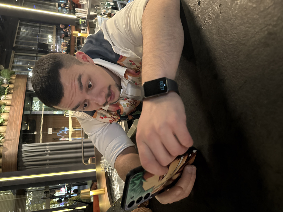 A male individual, approximately late 20s to early 30s, with short dark hair and a mustache, is leaning over a dark bar counter. He wears a white collared shirt with a colorful, floral-patterned vest-like garment. His eyes are wide and mouth slightly ajar, indicating a focused or surprised expression directed at a smartphone he holds horizontally with both hands. The phone features a case depicting an anime-style character. A dark smartwatch, displaying "00:27," is visible on his left wrist. The background shows a contemporary, dimly lit bar interior, identified as The Martini Bar at Graffit Gallery Design Hotel in Varna, Bulgaria. Visible elements include shelves with glassware and liquor bottles, decorative plants in woven baskets suspended above, dark curtains, and warm ambient lighting, with blurred figures and reflections discernible through distant glass partitions.