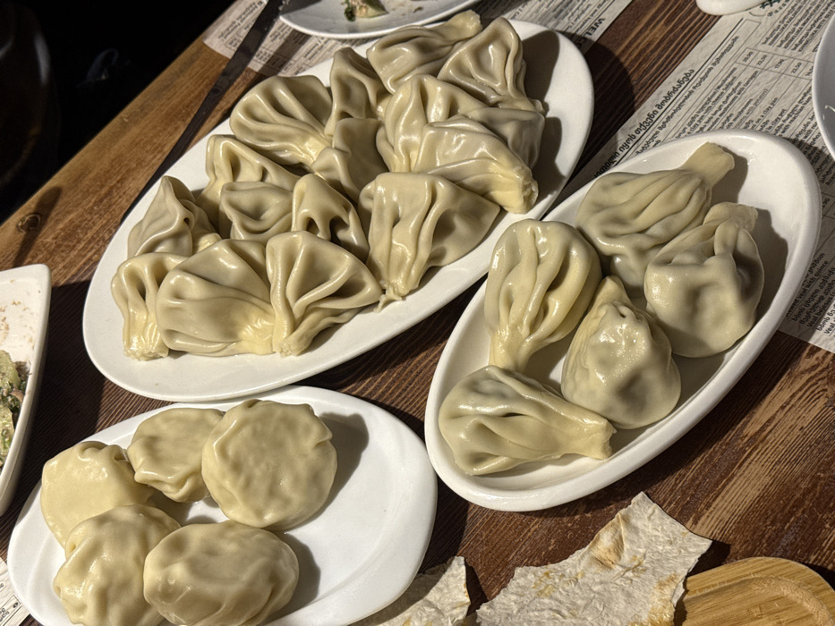 The image displays a close-up, elevated view of multiple plates of Khinkali (Georgian dumplings) arranged on a dark brown wooden table within what appears to be a restaurant setting in Tbilisi, Georgia.
Three white plates of varying shapes are prominent. The leftmost plate is oval, holding approximately six smooth, round Khinkali. The largest plate, centrally located and rectangular, contains numerous Khinkali, estimated at fifteen to twenty, characterized by their traditional pleated tops and conical shapes. A smaller, oval plate in the bottom-center also holds about six Khinkali, predominantly with pleated tops.
The wooden table surface exhibits a visible grain. Partially visible paper placemats or menus with Georgian script and numerical prices are situated beneath some of the plates, confirming the cultural context. To the bottom-left, a crumpled piece of light-colored flatbread rests on a small, light-colored wooden cutting board or trivet. A portion of another white plate with remnants of food, possibly a salad, is visible in the top-left corner. The lighting is indirect, suggesting an indoor environment. No individuals are visible in the frame.