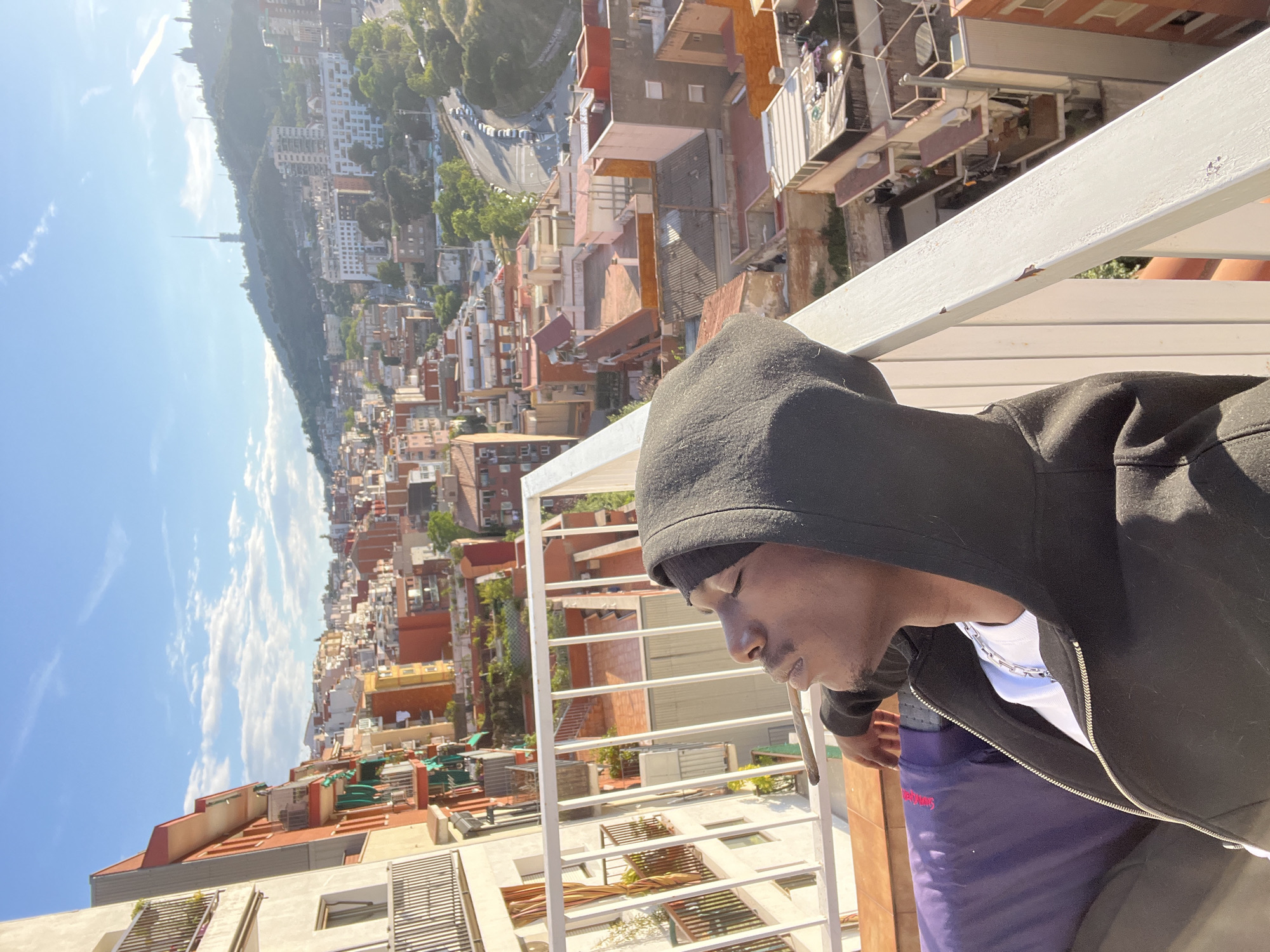 A person in a black hoodie stands on a balcony overlooking a cityscape. The backdrop is a dense urban area with buildings of various heights and colors, primarily orange and white, extending towards a green, tree-covered hill. The sky is bright blue with scattered clouds. The person appears to be looking down or introspective. The balcony has white railings, adding to the composition's depth. This is Barcelona, Spain.