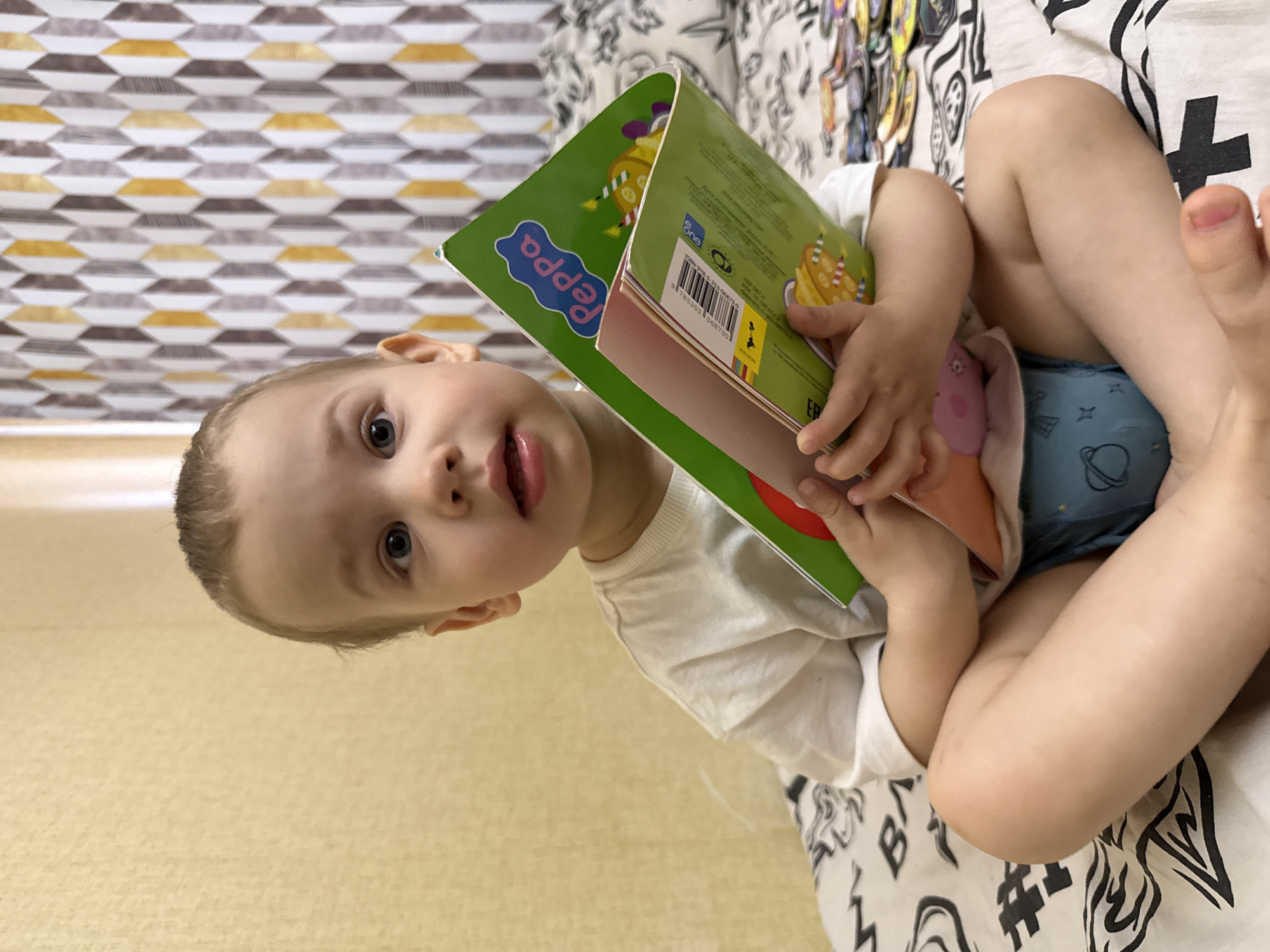 A young child is sitting on a patterned bed or couch, holding a green and red children's book with "Peppa" written on the cover. The child is wearing a white t-shirt and blue underwear with a pattern of planets and stars. The background includes a geometric patterned surface behind the child's head, and the surface beneath the child appears to be a solid color. The location is the city of Unknown, Unknown.