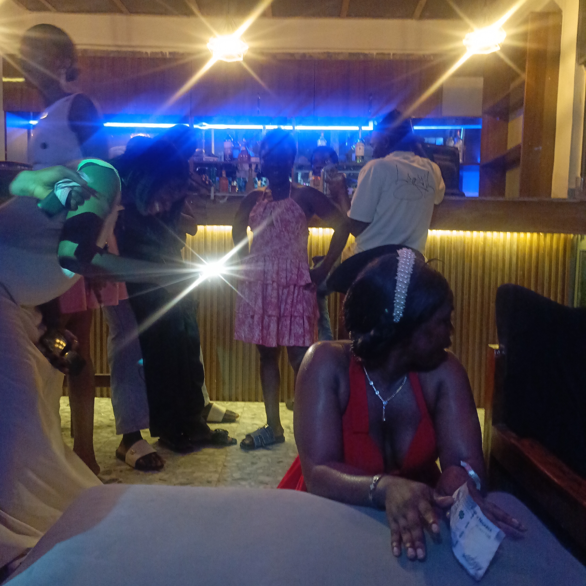 The image depicts a nighttime scene inside a bar or lounge in Akwanga, Nigeria. The location is illuminated by warm overhead lighting, complemented by blue accent lighting behind the bar. Several individuals are present: A woman in a red dress sits in the foreground holding Nigerian Naira currency. Other people are standing around the bar, including one in a pink dress and another in a white t-shirt, possibly interacting with the bartender. Drinks are visible on the bar. Some individuals are in motion. The atmosphere appears to be lively and social.